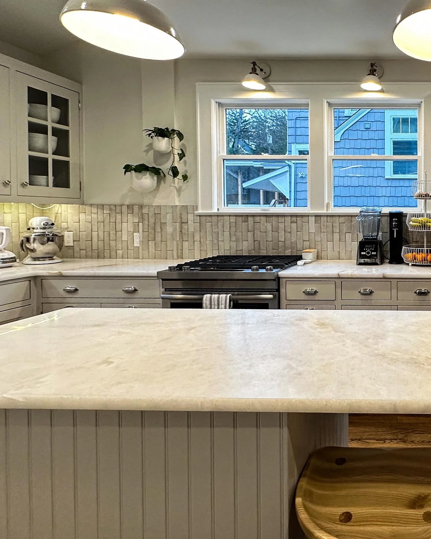 A spacious kitchen with beige cabinetry, a marble countertop, a stove, and a large window with three sections showing a blue house outside. There are potted plants on the wall, a blender, a coffee maker, and a fruit basket on the counter.