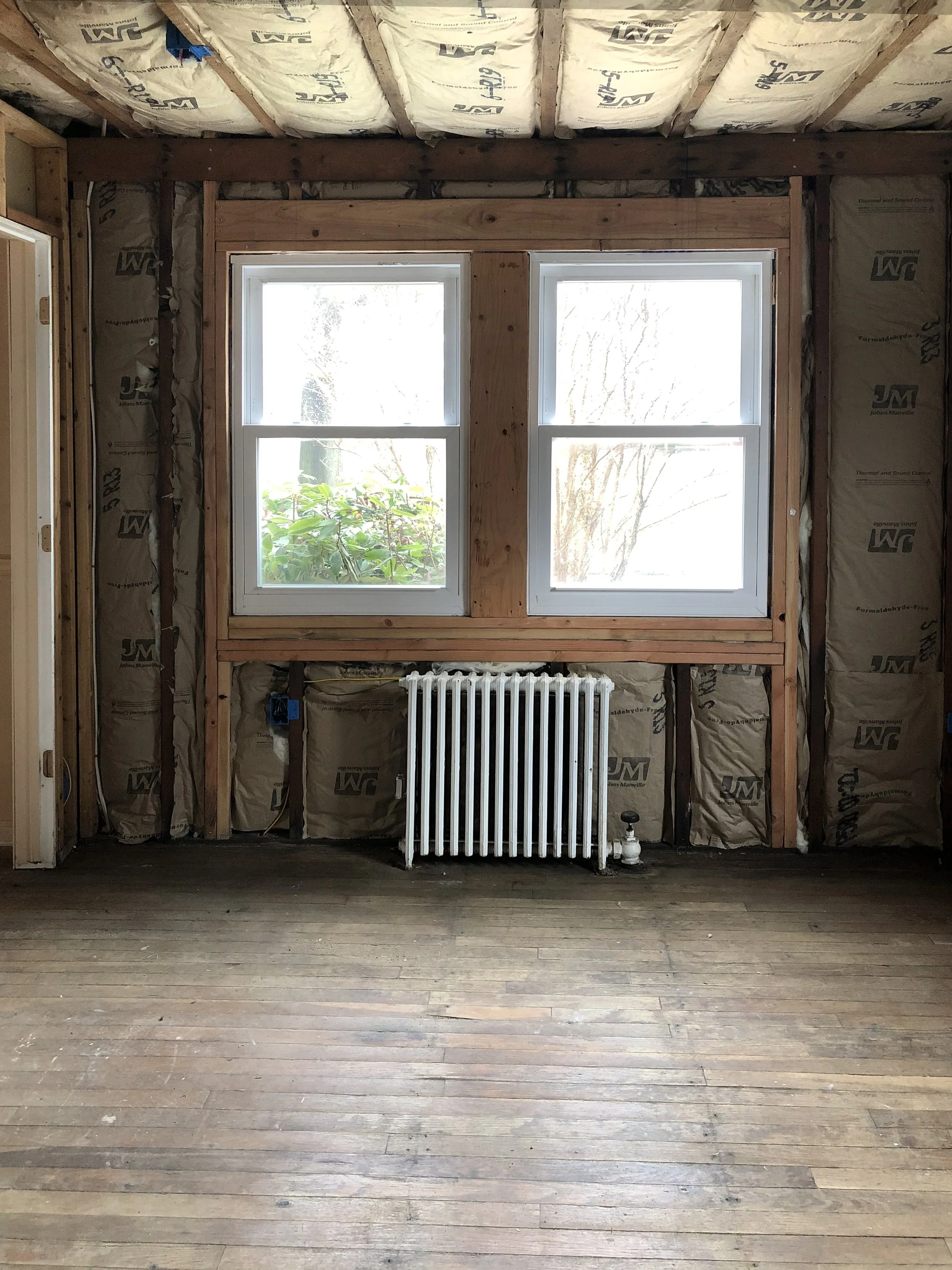 Interior of a room under construction with two rectangular double-pane windows, a white radiator below the windows, unfinished insulation on the walls and ceiling, and a wooden floor.