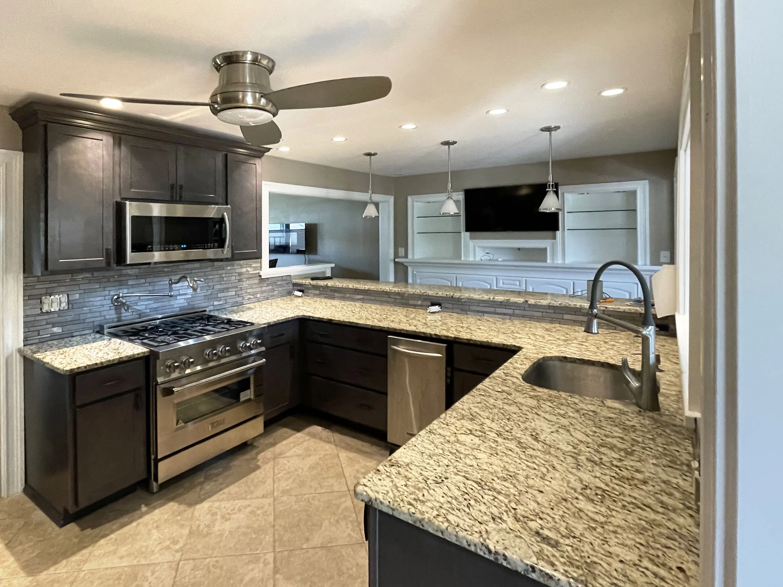 Kitchen with dark wood cabinets, granite countertops, stainless steel appliances, a ceiling fan, and a view into a living room with a wall-mounted TV.