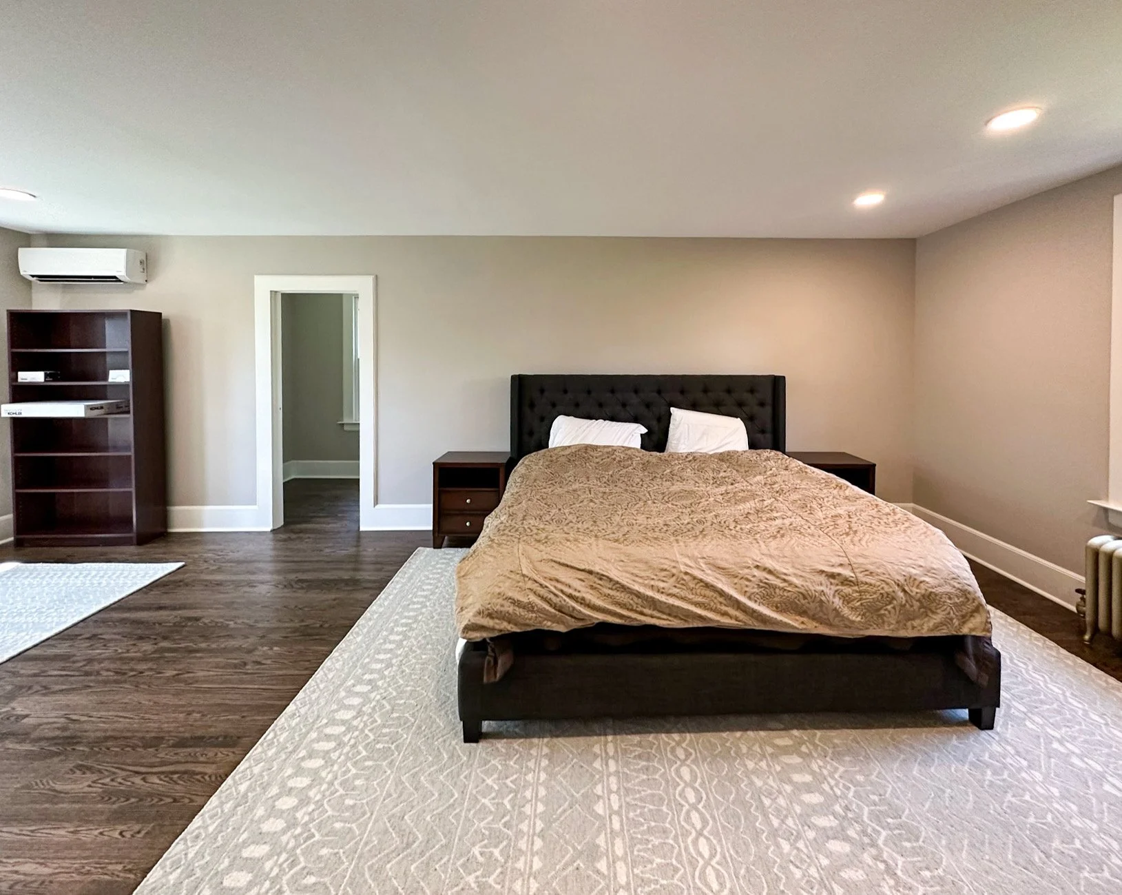 A spacious bedroom with a large bed and a black tufted headboard, flanked by two nightstands, on a patterned beige rug. To the left, there is a wooden bookshelf and an air conditioning unit on the wall. The room has hardwood floors, beige walls, and 