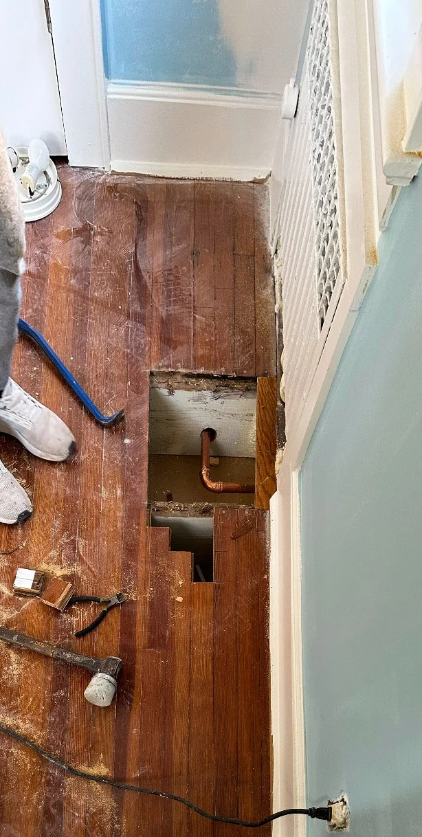 A section of hardwood floor removed to reveal plumbing pipes beneath. Tools and a person's foot are partially visible around the opening.