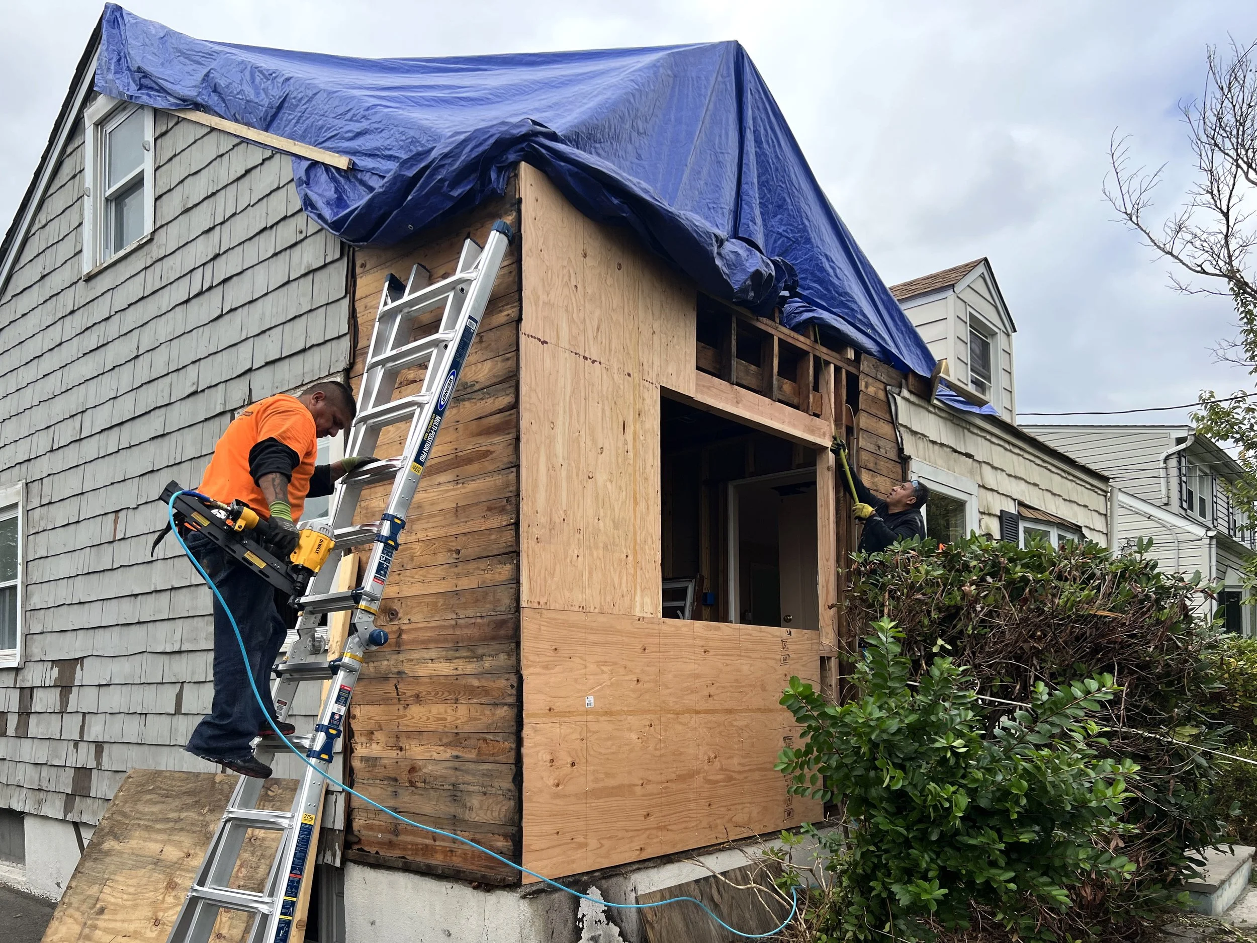 Workers are renovating a house, with one on a ladder and another working on the upper wall, as they attach wooden panels under a blue tarp covering the roof.