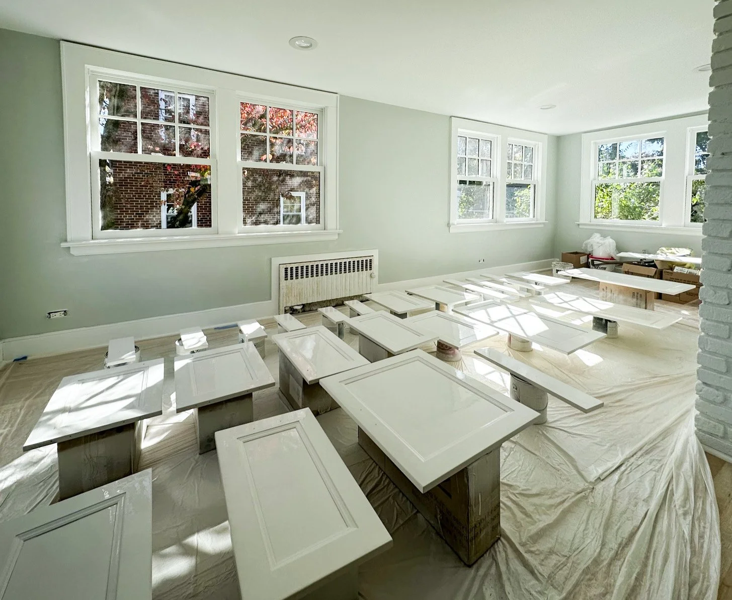 A bright room under renovation with white painted cabinet doors laid out on the floor and wrapped in protective paper. Large windows let in natural light, showing a view of trees and a brick building outside.
