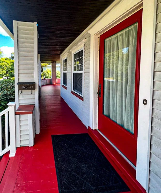 Front porch of a house with a red door with glass panel and curtain, white siding, black doormat, and a mailbox marked 'MAIL 102'