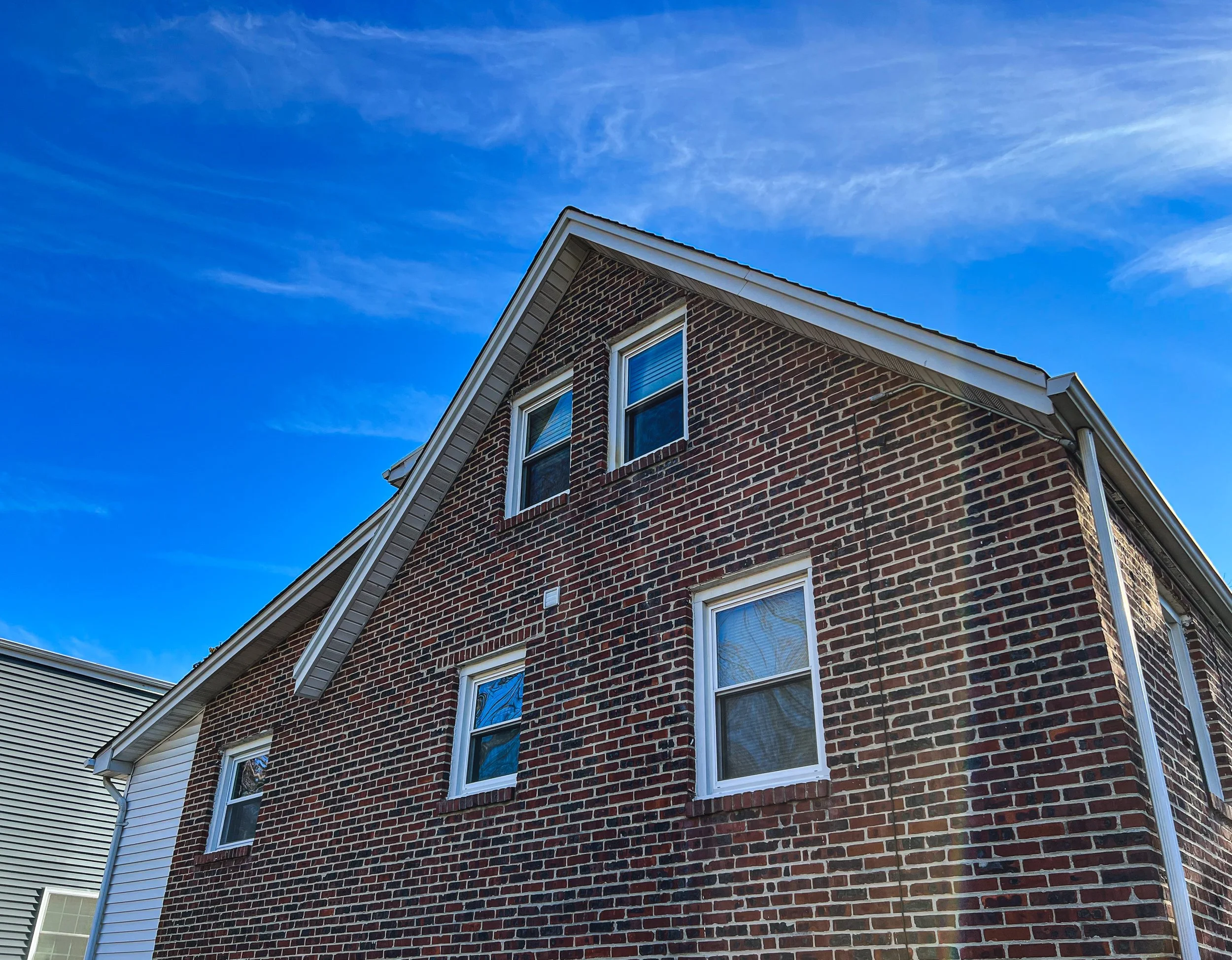 A brick house with a sloped roof and four windows against a blue sky with some clouds.
