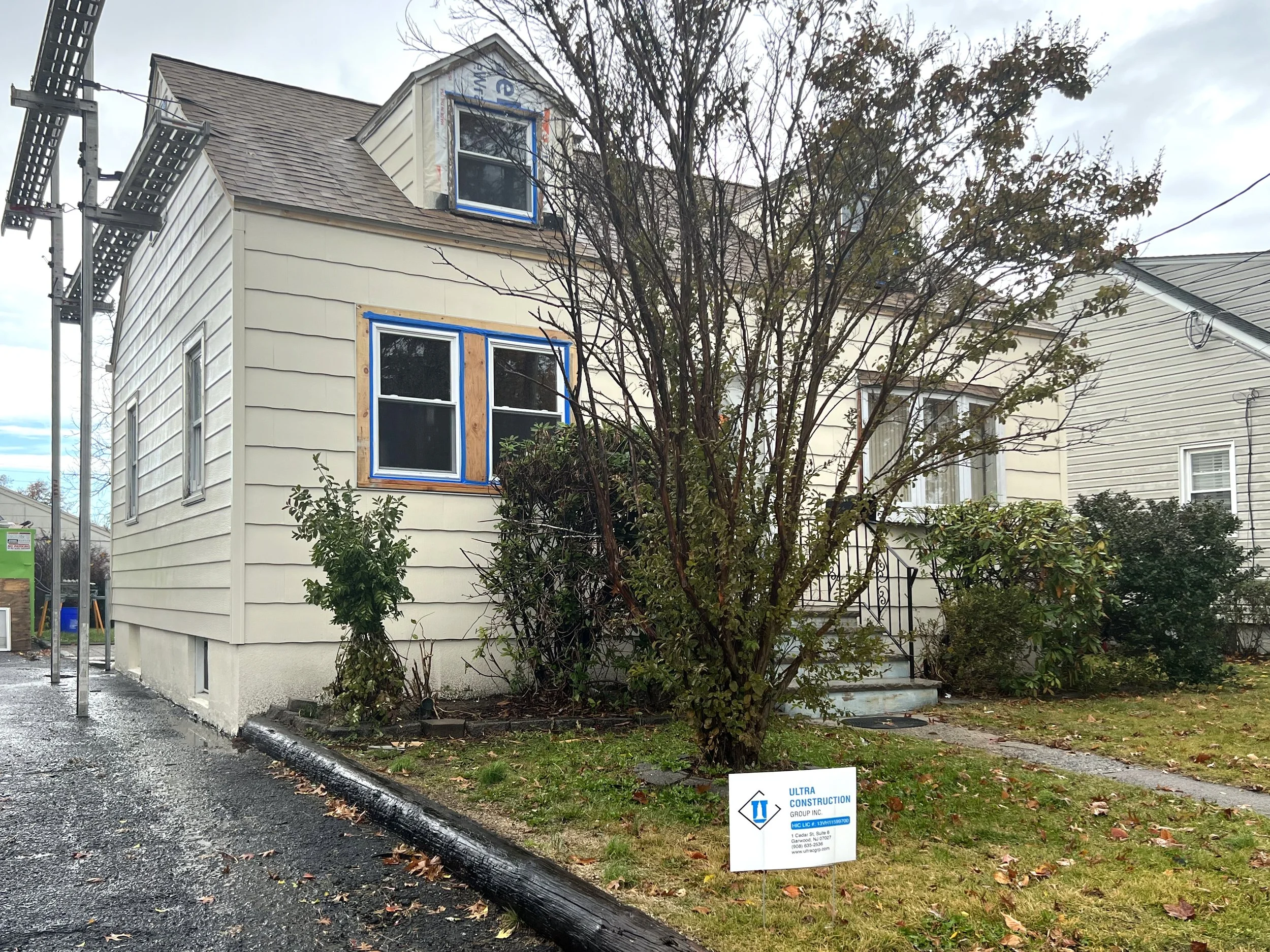 A house under renovation with new window trim, surrounded by bushes and a tree, next to a construction sign, on a wet road and grassy yard.