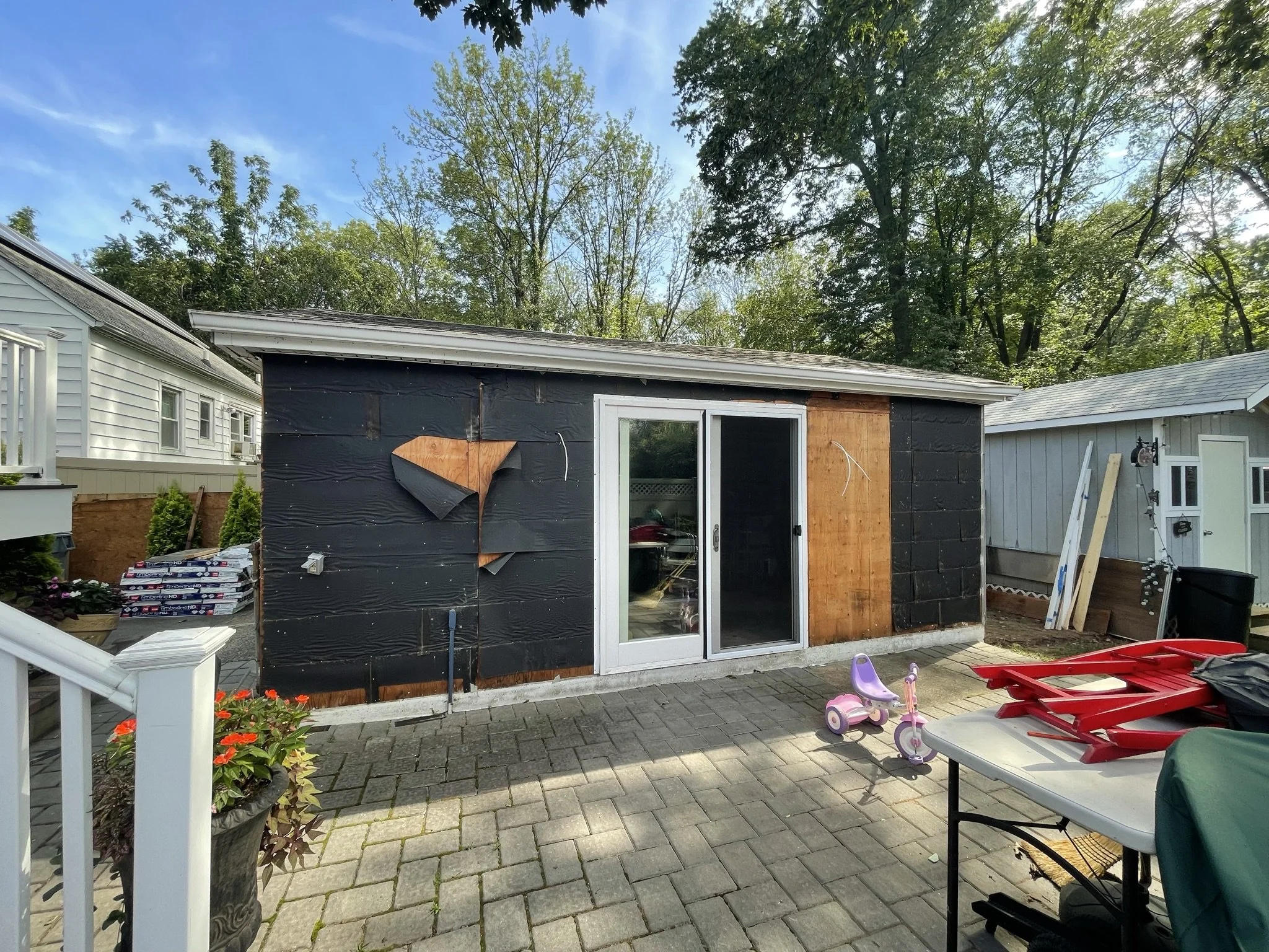 Backyard scene during construction showing a small building with black and wooden exterior, a sliding glass door, and surrounded by trees. There are children's toys, a table with red chairs stacked, and construction materials nearby.