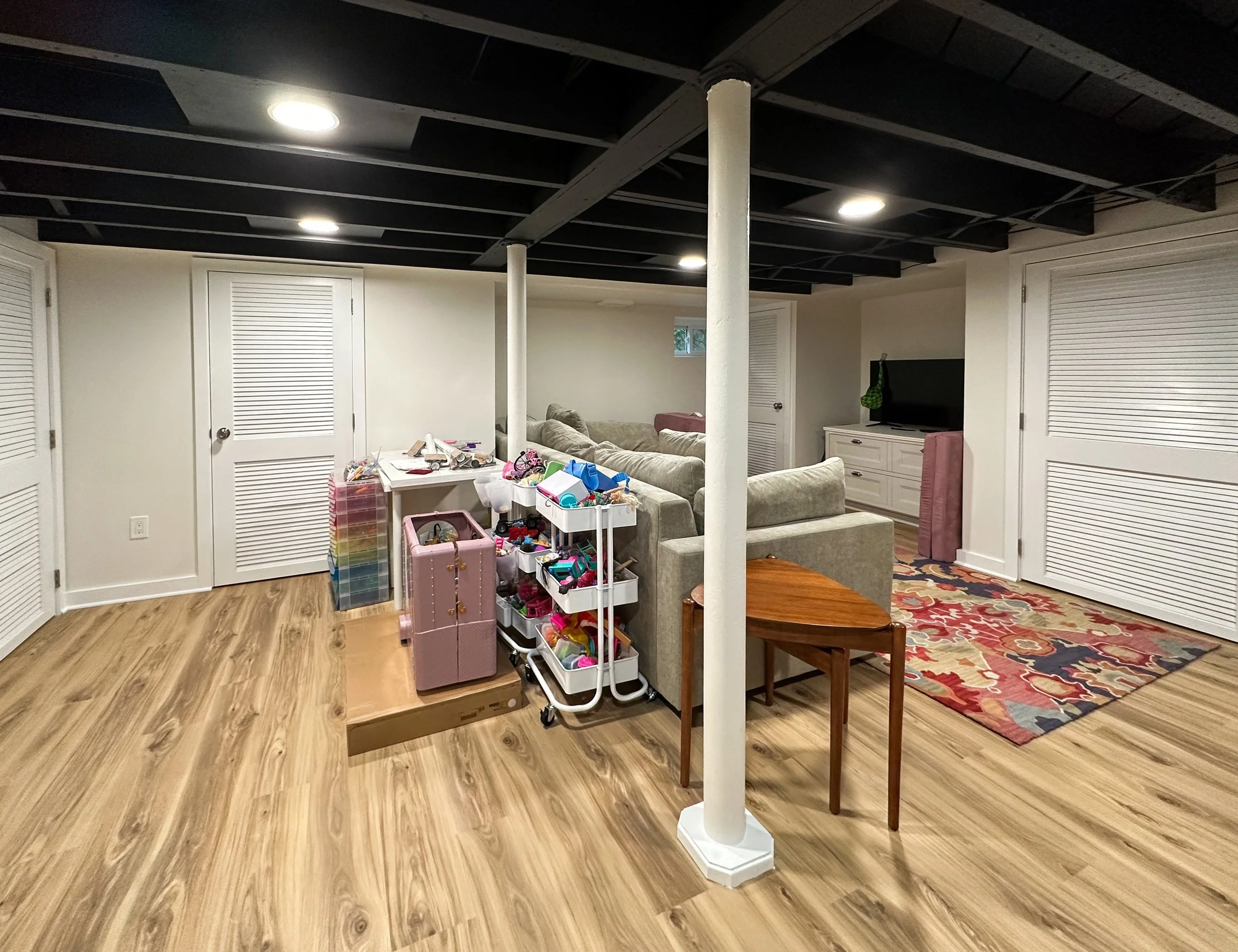A basement living space with light wood flooring, white walls, and a black ceiling with recessed lighting. There are white louvered closet doors on the walls. A gray sectional sofa is in the center of the room, with a small wooden side table nearby. 