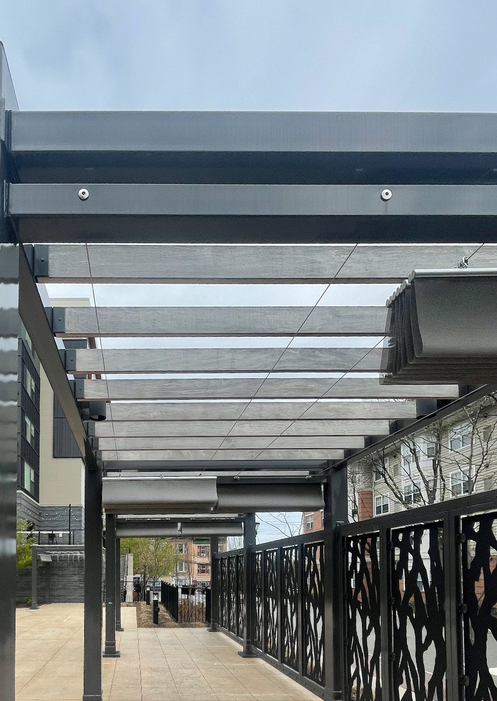 Covered outdoor walkway with metal beams and a decorative black metal railing on the right side, with a cloudy sky overhead.