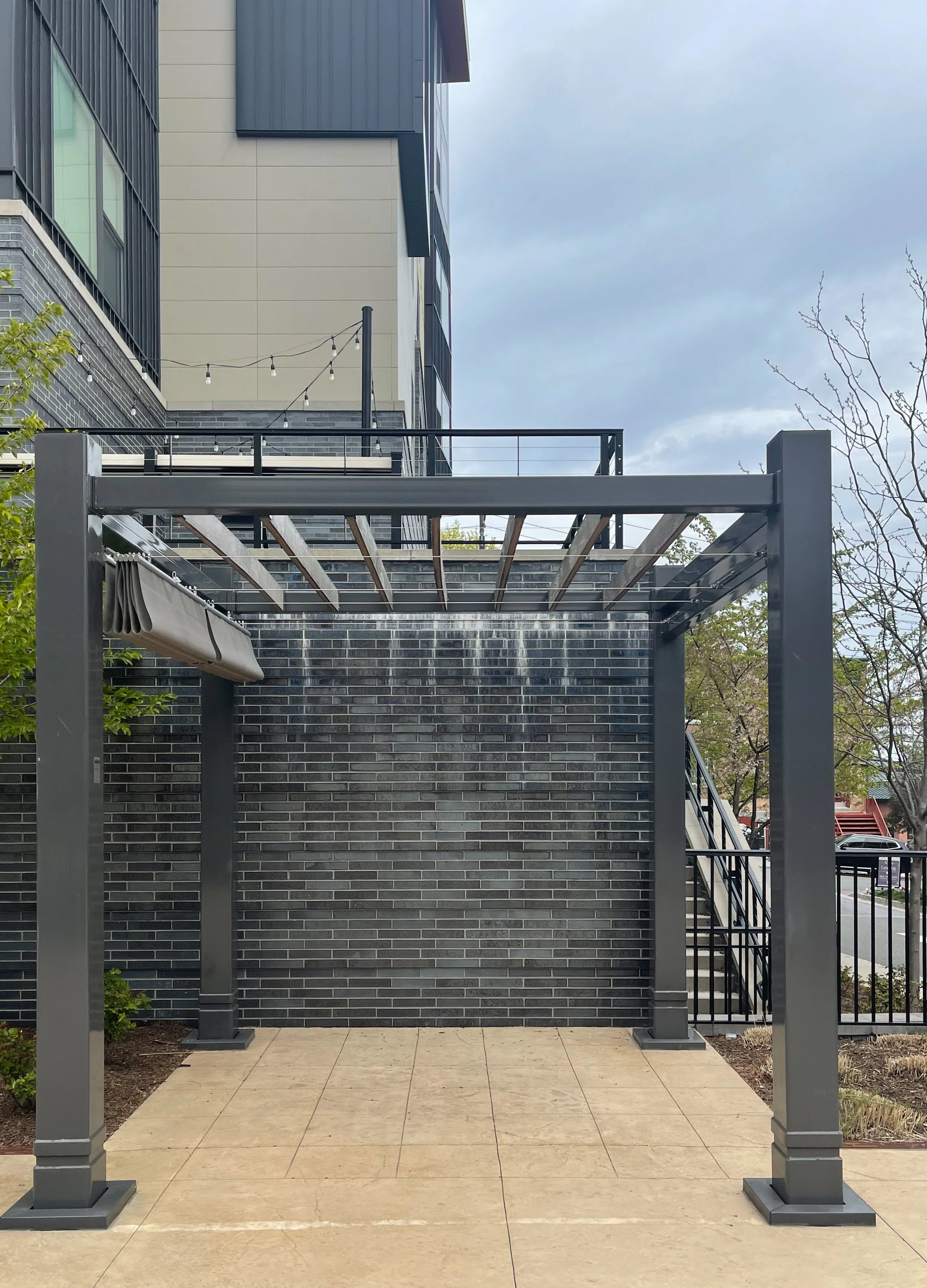 Outdoor patio area with a metal pergola, brick wall with water dripping from an overhead water feature, stairs on the right, and a building with balconies in the background.