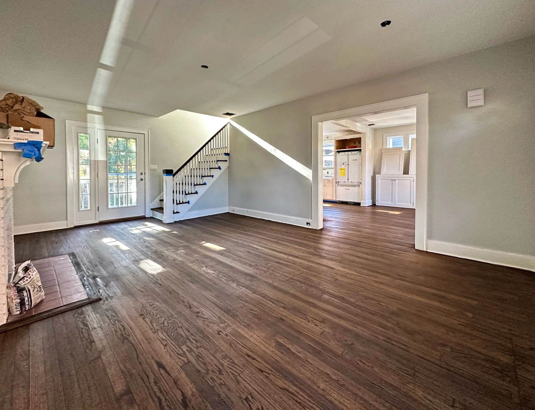 Empty living room with hardwood floors, a staircase, a door with glass panels leading outside, and an adjacent open kitchen space with white cabinetry.