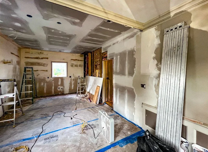 Room under renovation with drywall installation, ladders, construction materials, and a small window.