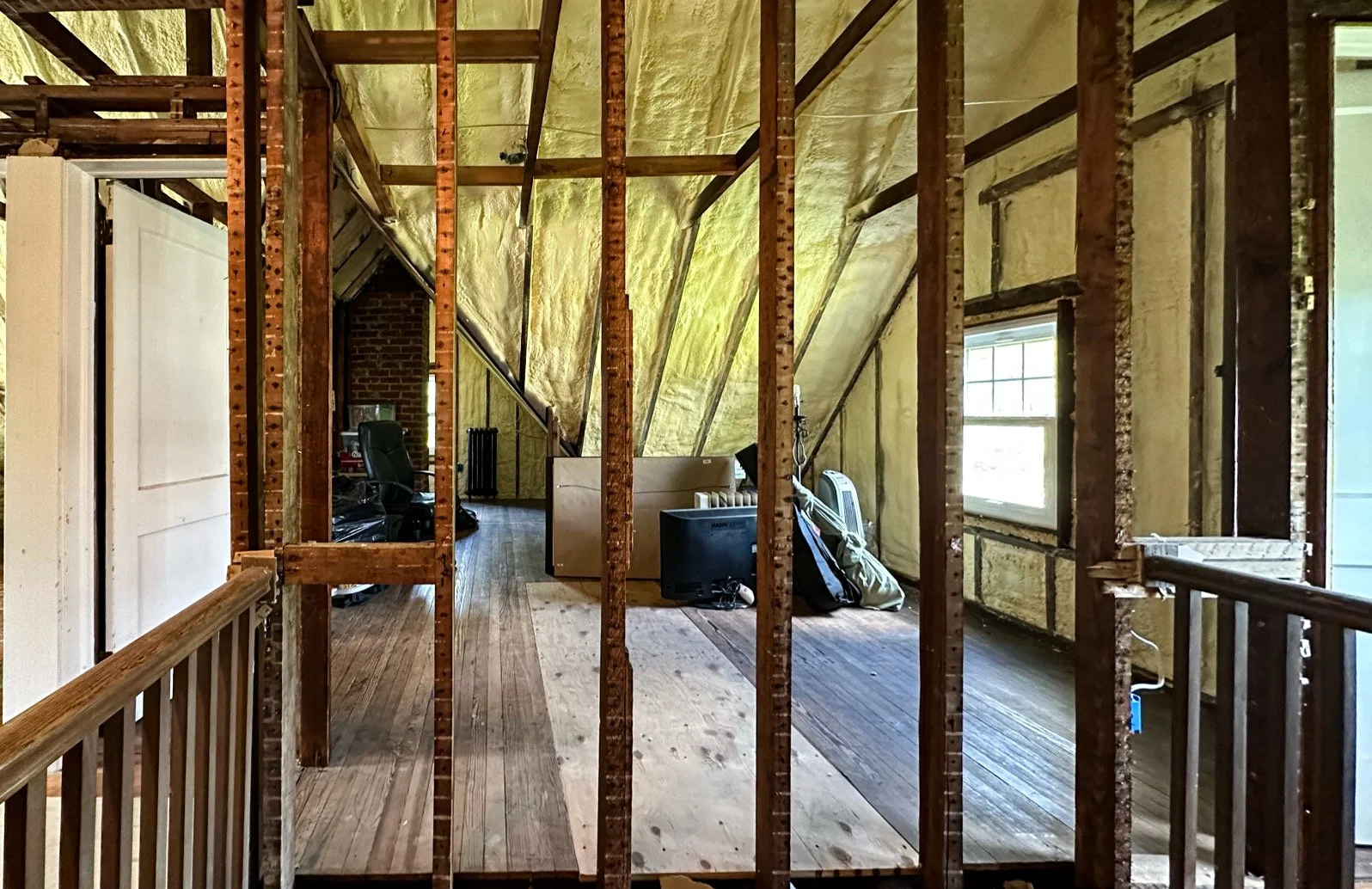 Interior of a house under renovation with exposed wall studs, insulation, and some furniture and boxes on the wooden floor.