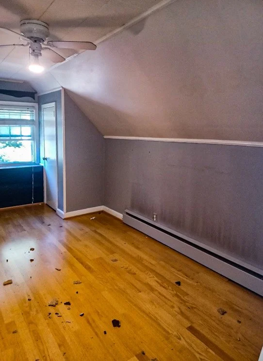 Empty room with hardwood floors, gray walls, a ceiling fan, a window with blinds, and debris scattered on the floor.