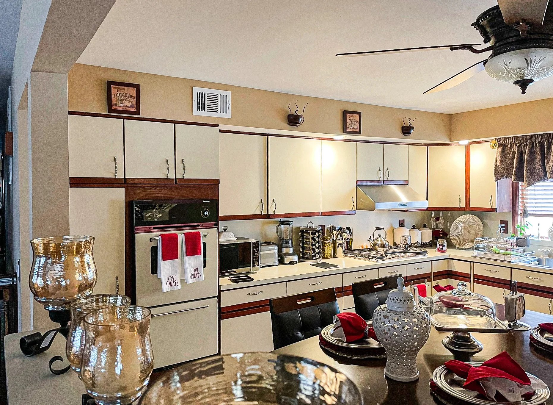 A kitchen with white cabinets, a beige wall, and various appliances including an oven, microwave, toaster, and blender. There is a dining table with red napkins, plates, and a decorative white vase. The kitchen has a ceiling fan and window with curta
