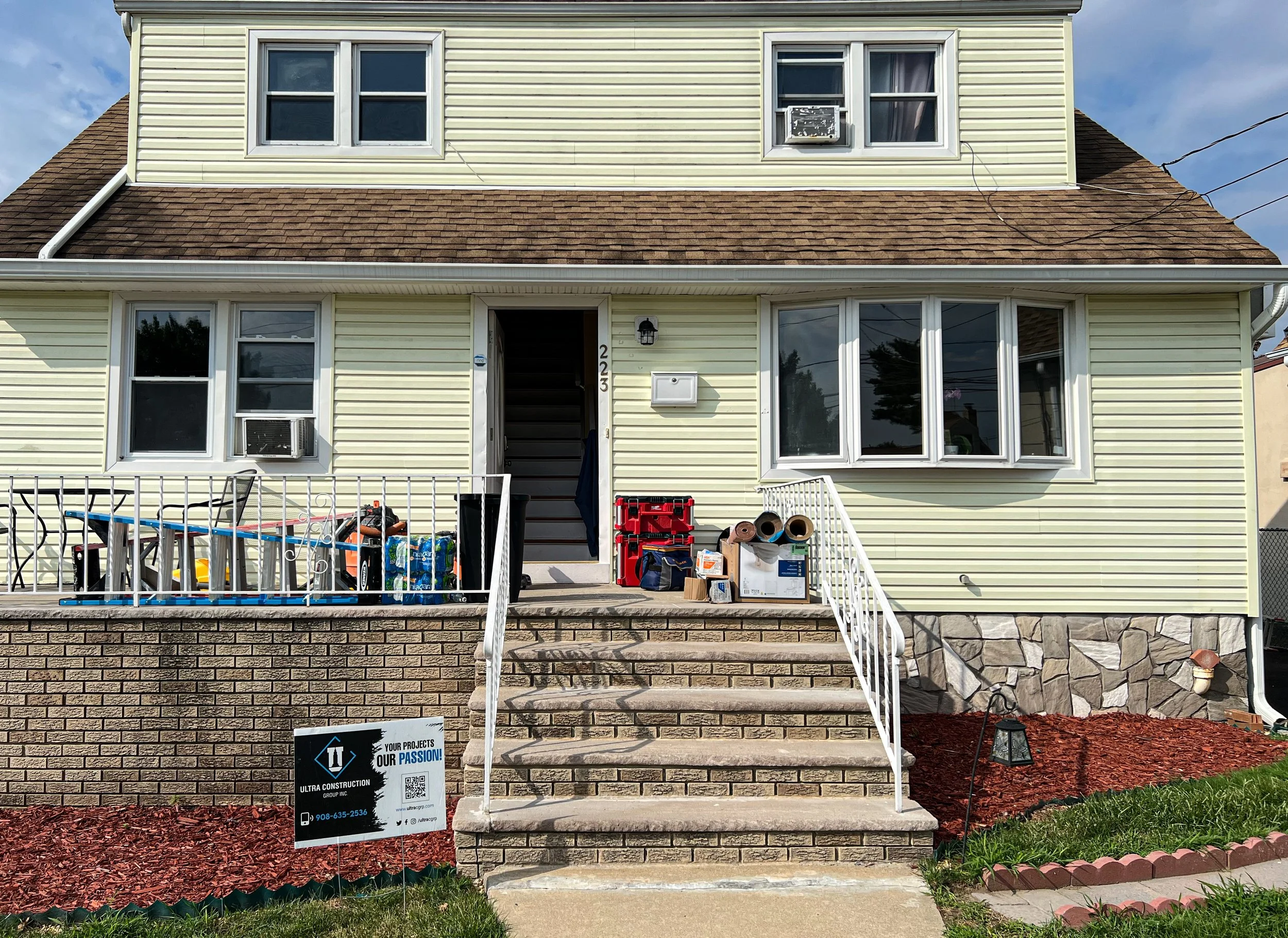 Front view of a yellow house with multiple windows, stairs leading to the entrance, and various construction materials and tools on the porch.
