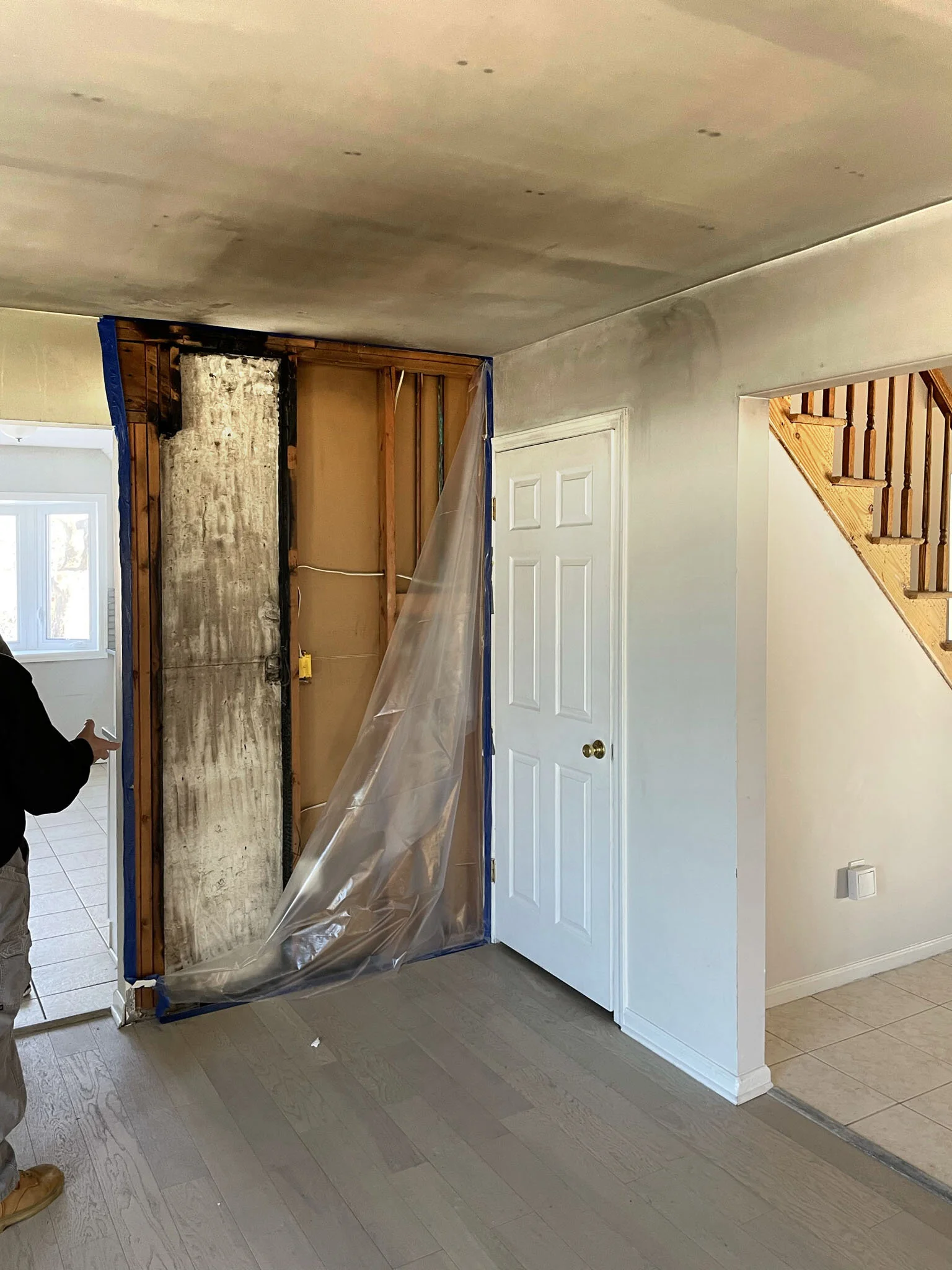 Interior wall renovation showing exposed framing and insulation, with plastic sheeting covering part of the wall, next to a doorway and staircase.