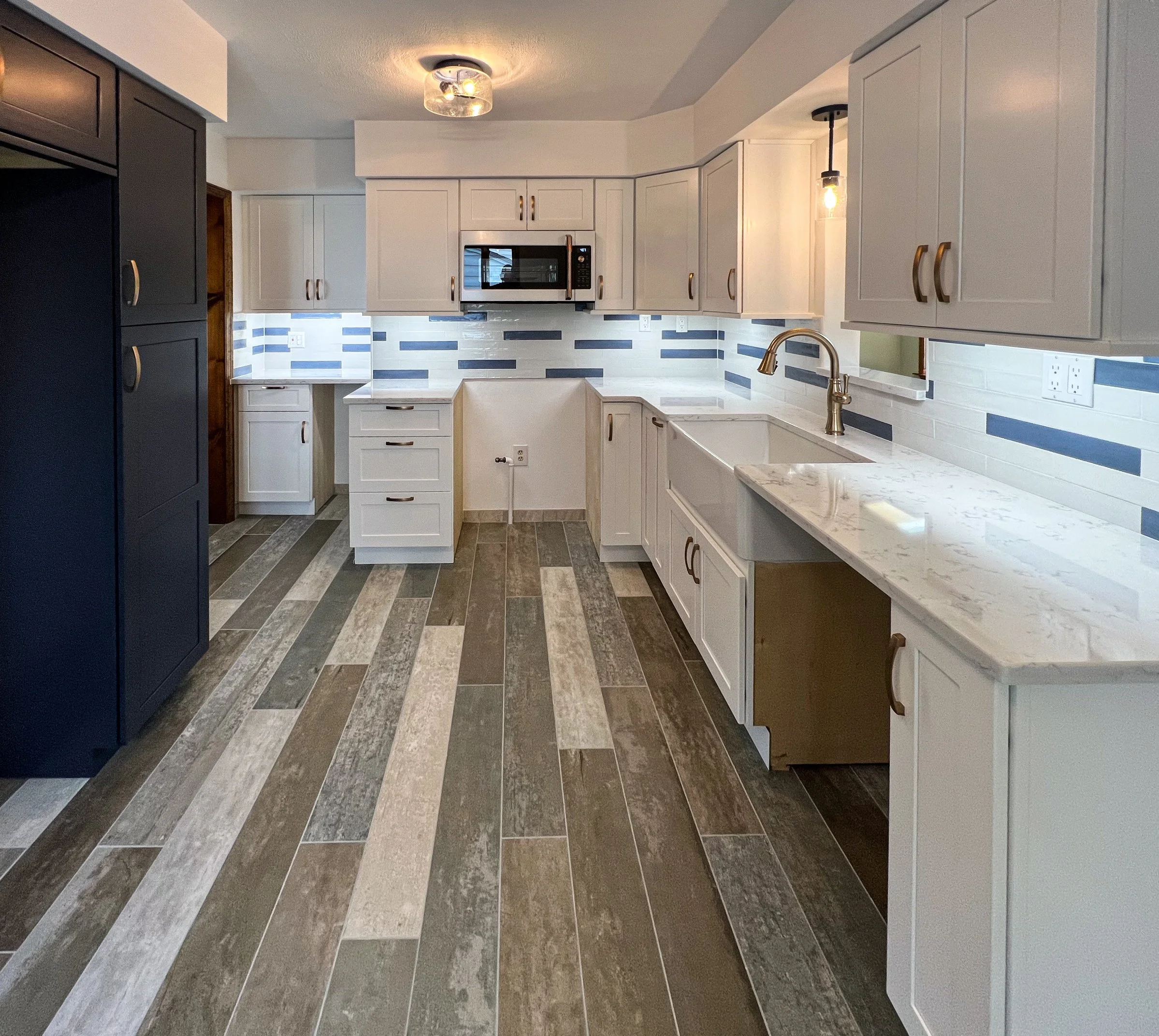 Modern kitchen with white cabinets, marble countertops, a white farmhouse sink, and a backsplash with blue and gray tiles. Wooden and dark-colored cabinets, and a multicolored wood-look tile floor.
