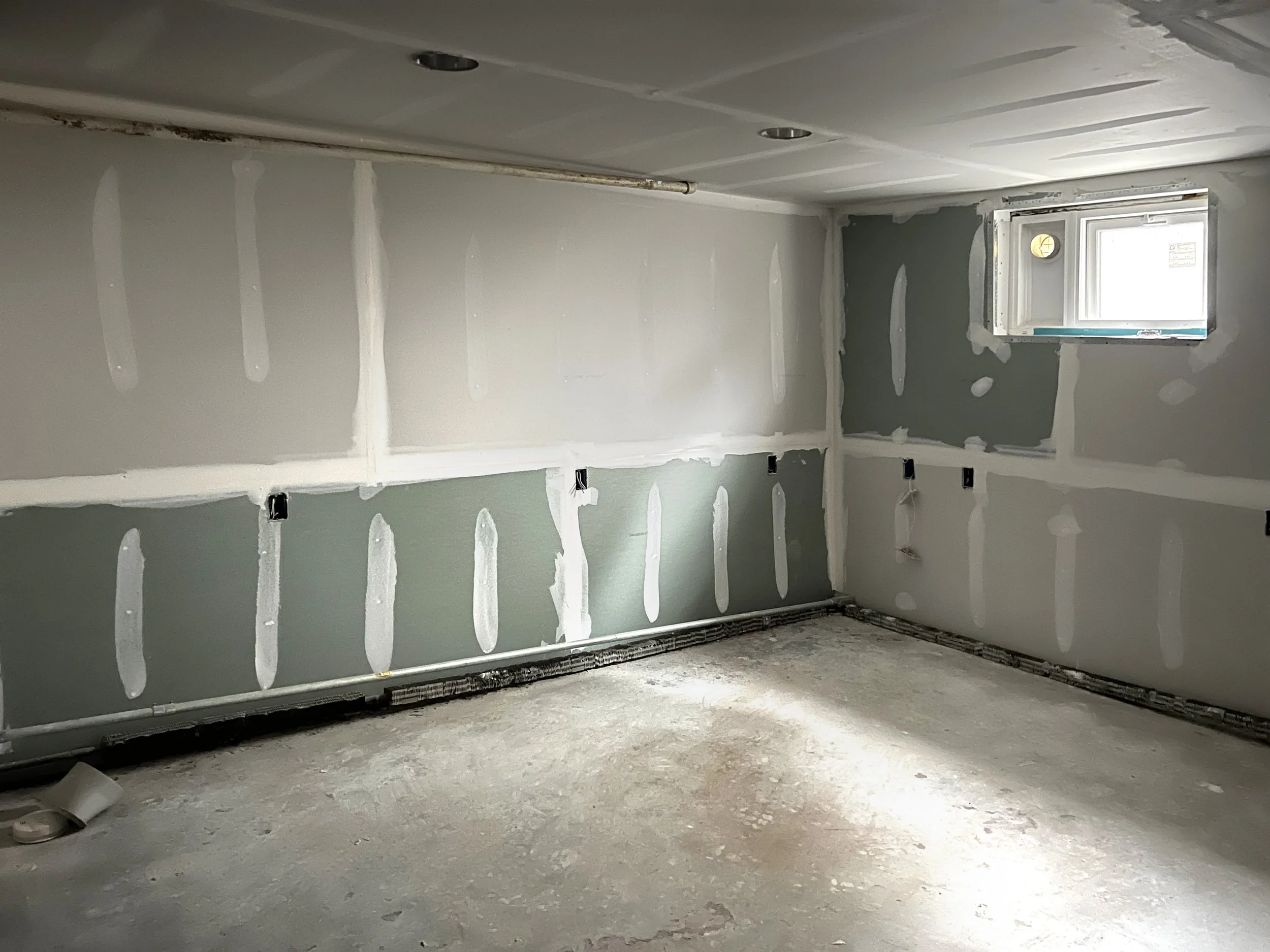 Unfinished room with drywall panels, some painted light green, others unpainted, with visible drywall seams and taped joints, and a small window in the corner.