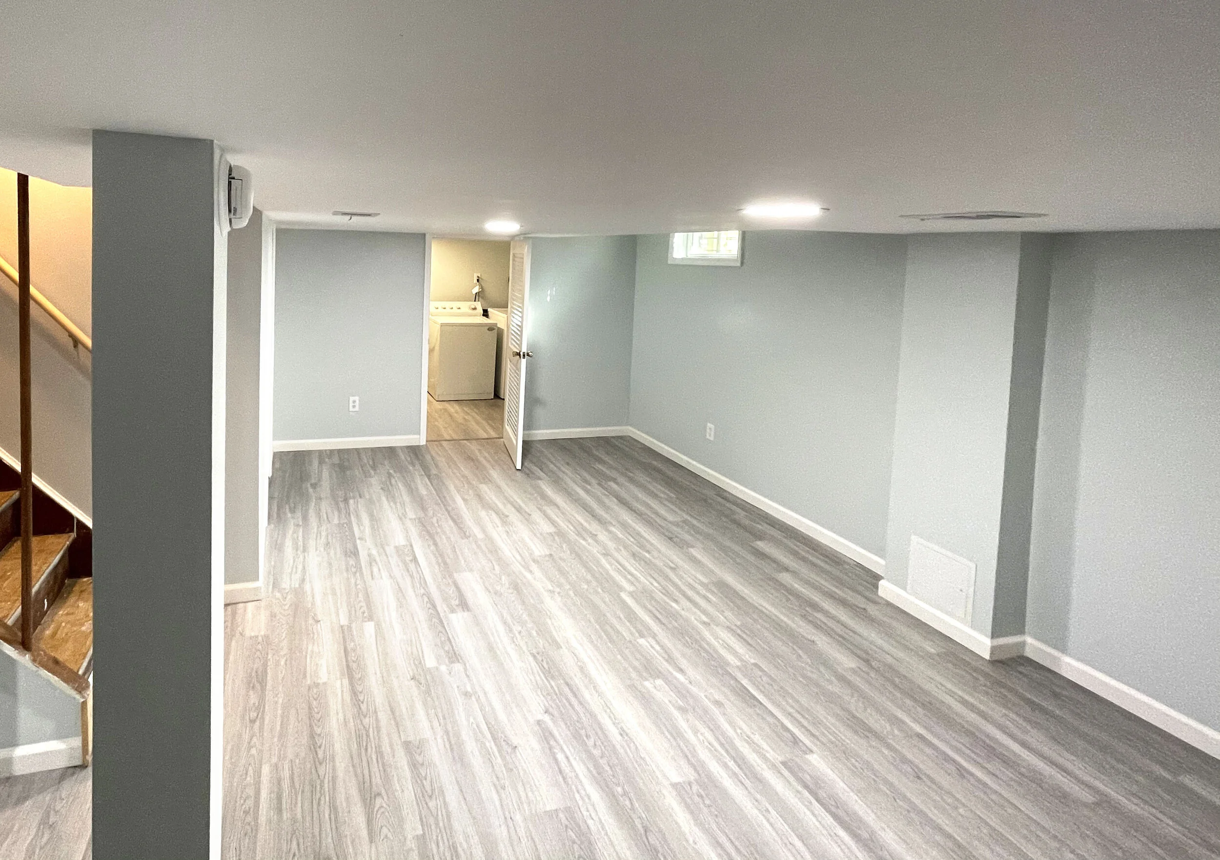 Empty room with light gray walls, gray wood flooring, a small window, and a laundry area with washer and dryer.