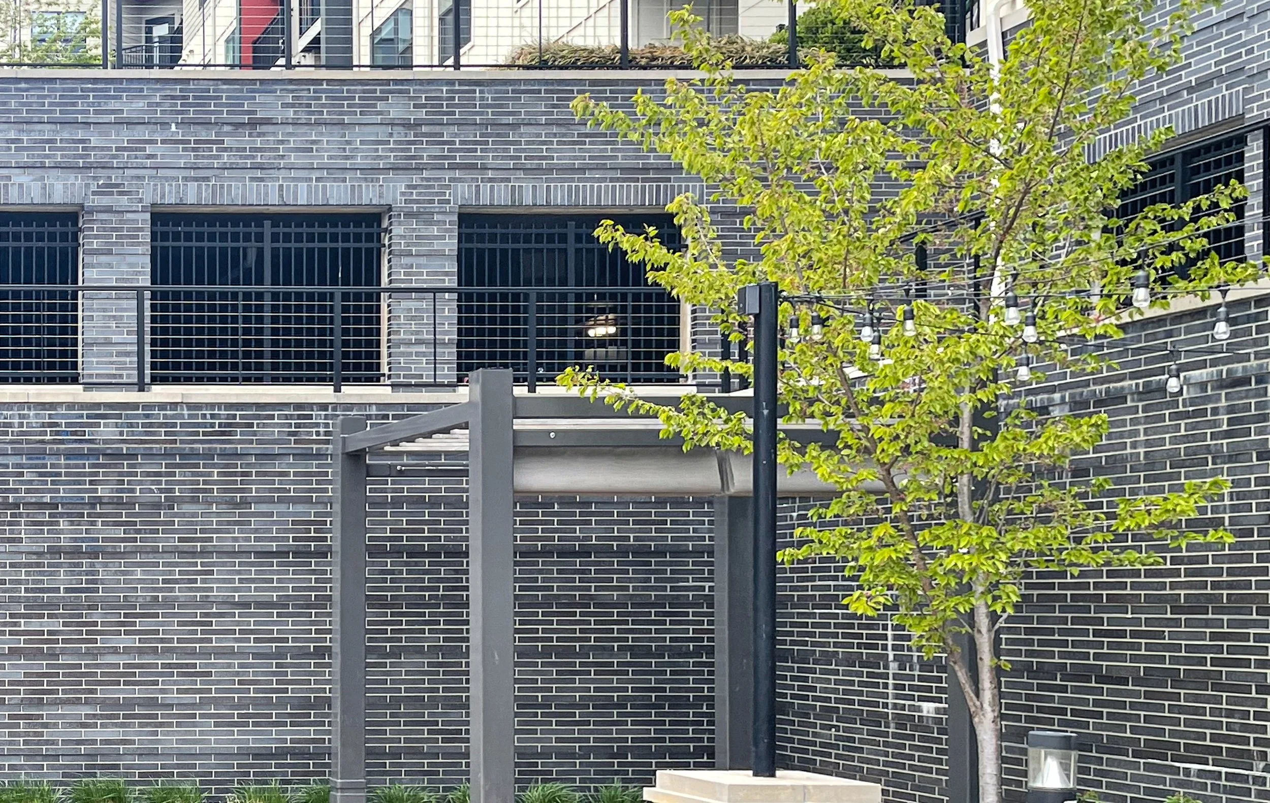 A modern urban scene with a brick building featuring black brick and metal railings, a small tree with green leaves, hanging string lights, and a metal structure in the foreground.