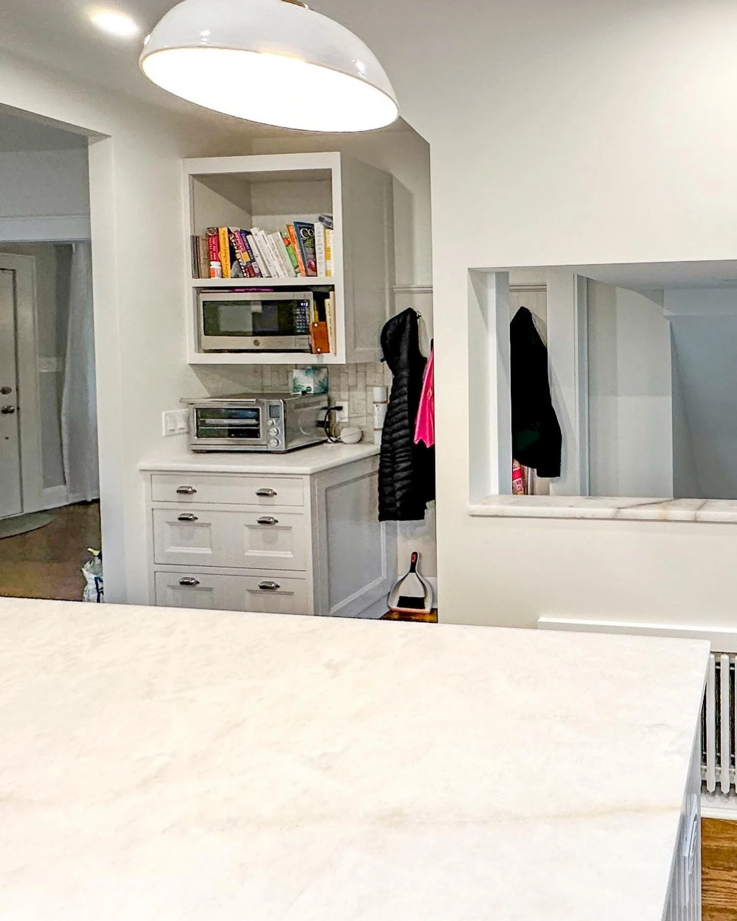 Kitchen with white cabinets, a countertop, a microwave, and a drawer cabinet. There are coats hanging on hooks and a dustpan on the floor. Part of a dining table or island is visible in the foreground.