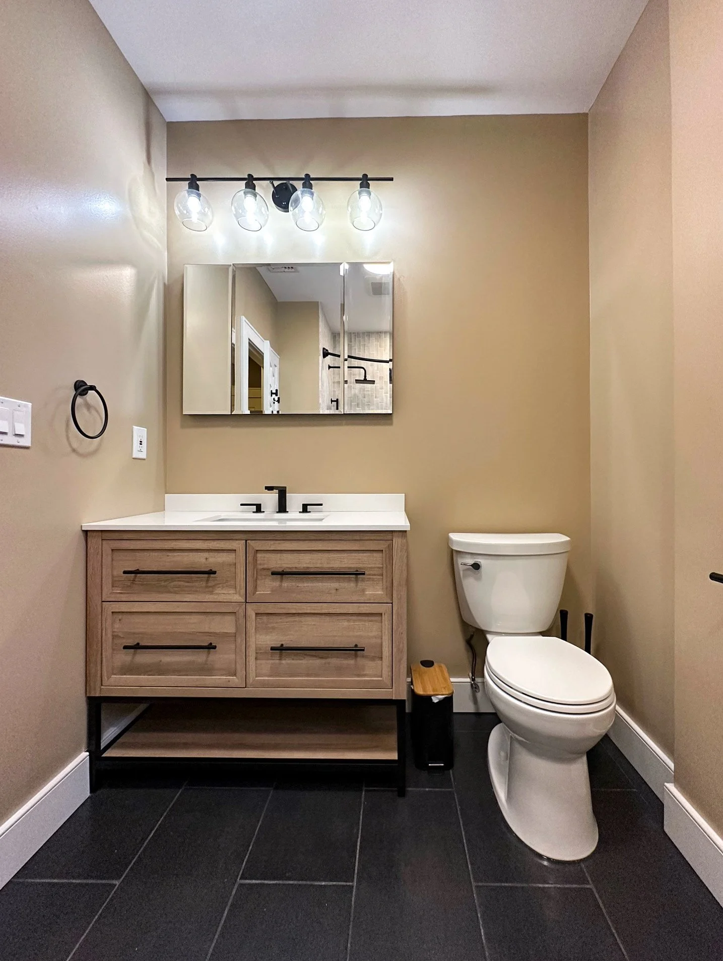 Modern bathroom with a wooden vanity, white countertop, black faucets, and a mirror. Light fixtures above the mirror, toilet on the right, black tile flooring.