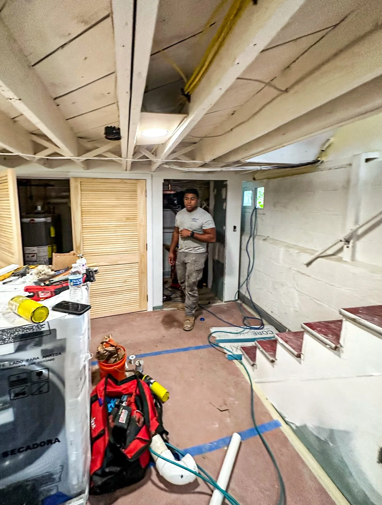 A man in gray work clothes standing in a basement under construction, with exposed ceiling beams and electrical wiring. Construction tools and materials are scattered around, including a red tool bag, water bottle, and a washer-dryer. The area has st