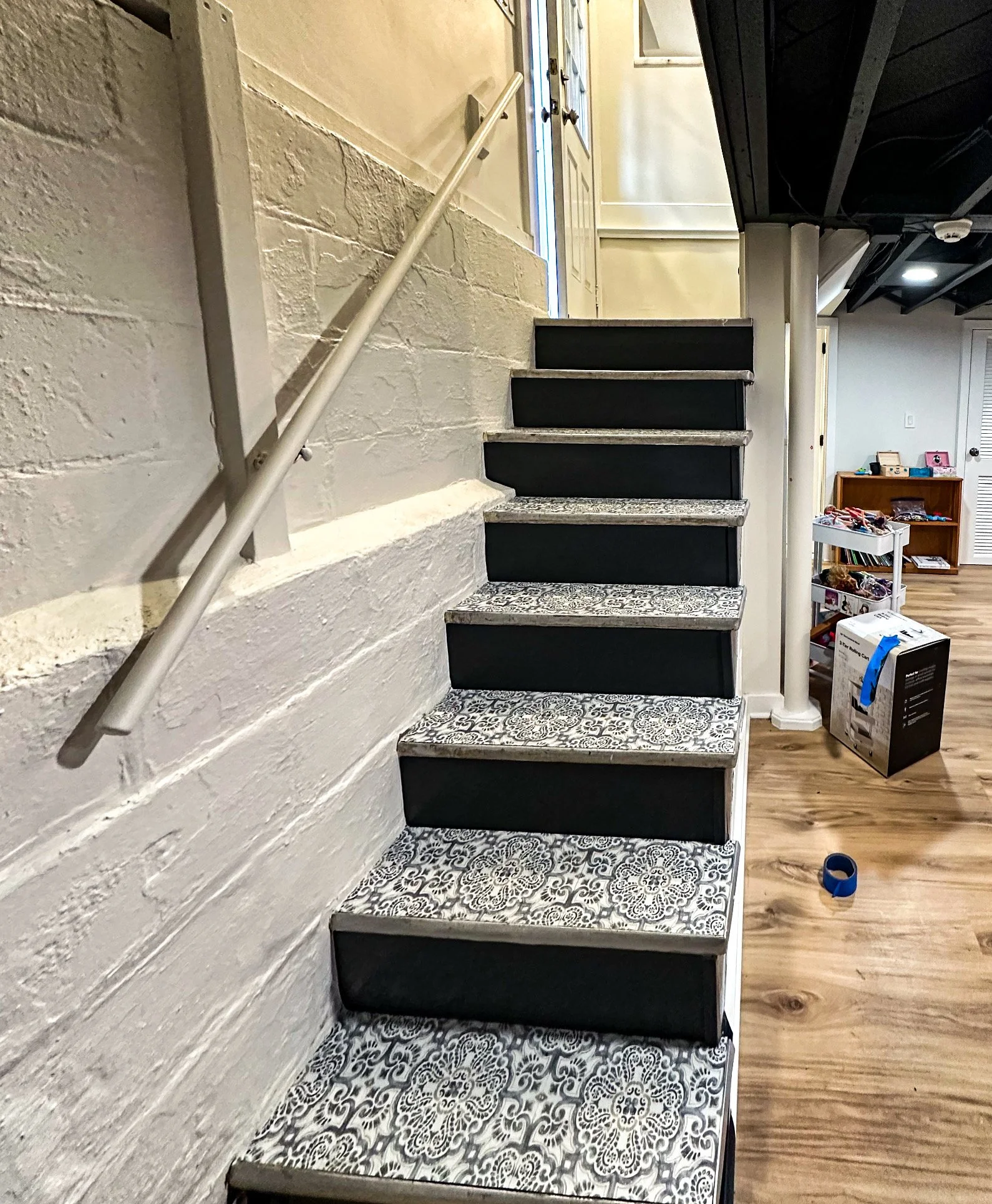 Indoor staircase with patterned black and white tiles on the steps, black risers, a gray handrail, and a white brick wall on the left side.