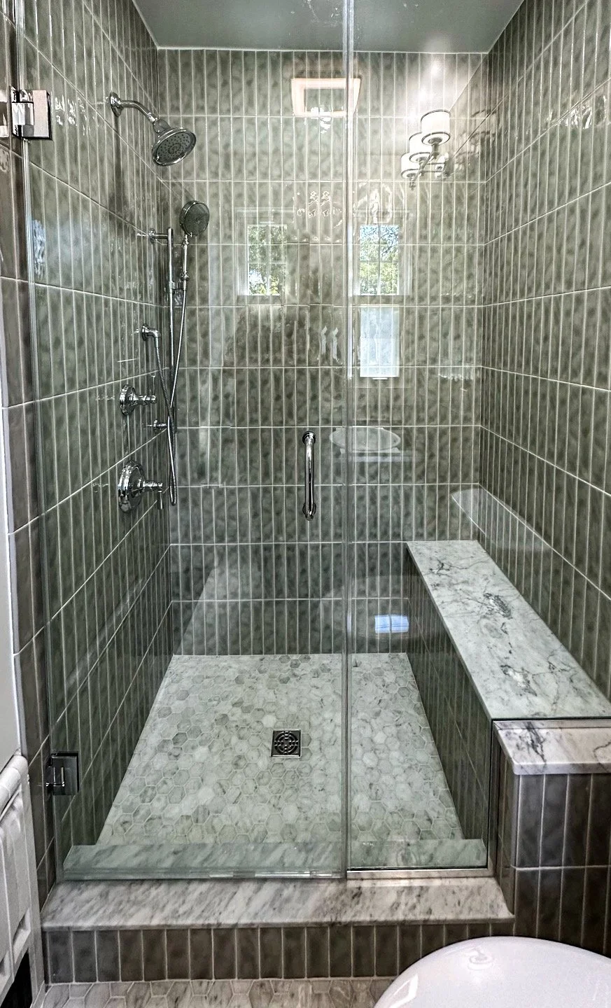 Modern walk-in shower with glass door, gray tile walls, mosaic floor tiles, a marble bench, and chrome fixtures, with natural light and small windows.