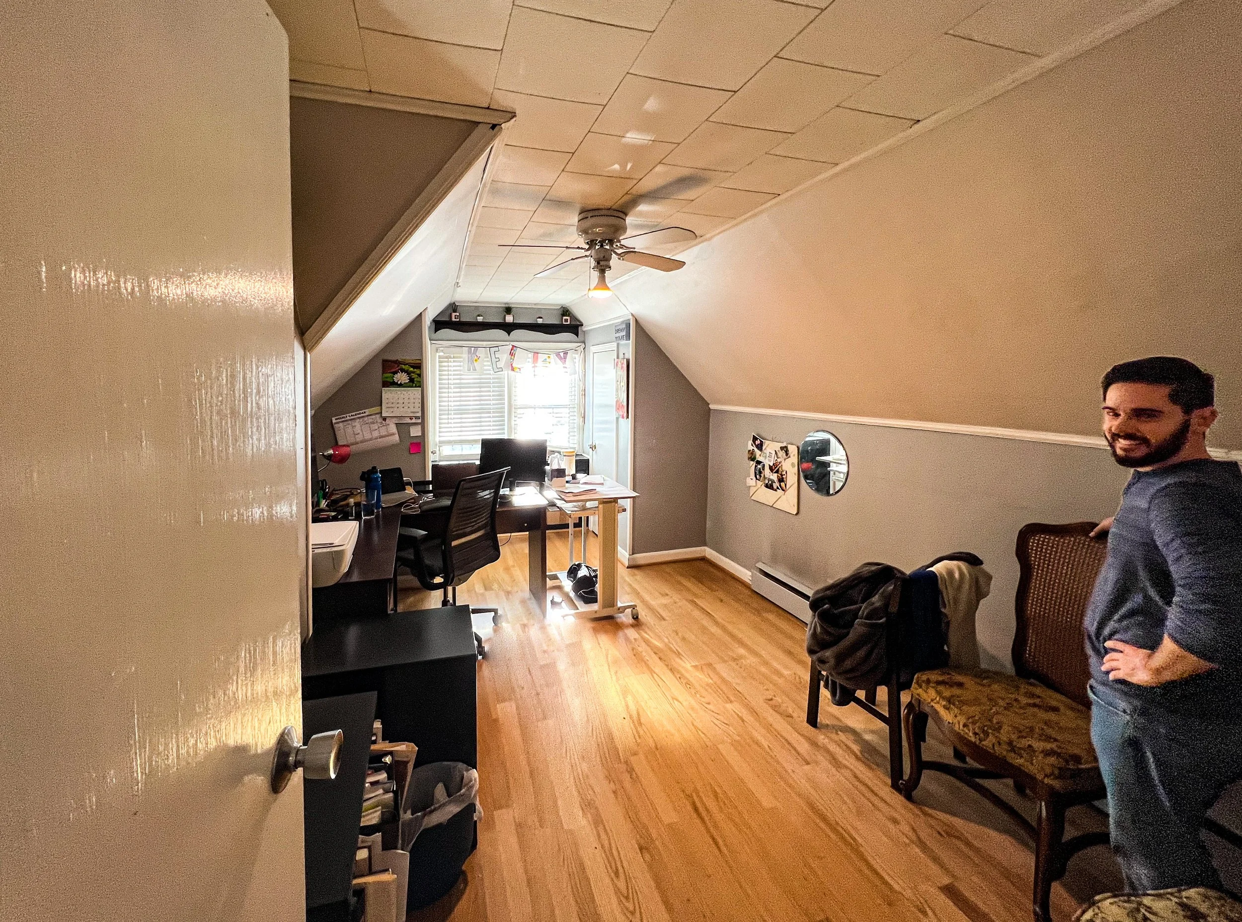A cozy attic home office with sloped ceiling, hardwood floors, desk with computer, chair, and a man smiling near a vintage chair.
