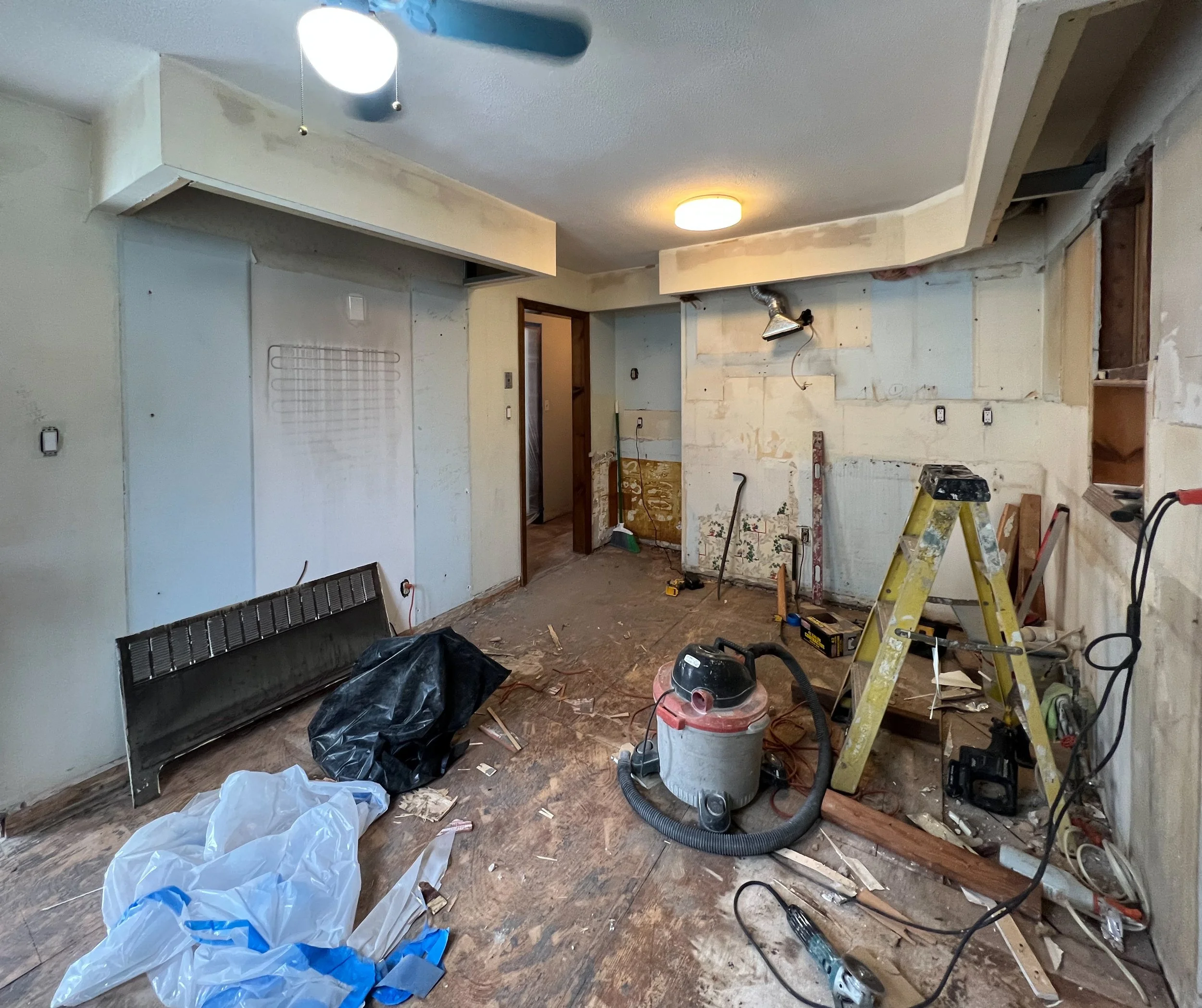 Room under renovation with tools and debris on the floor, including a vacuum, ladder, and construction materials, with unfinished walls and ceiling.