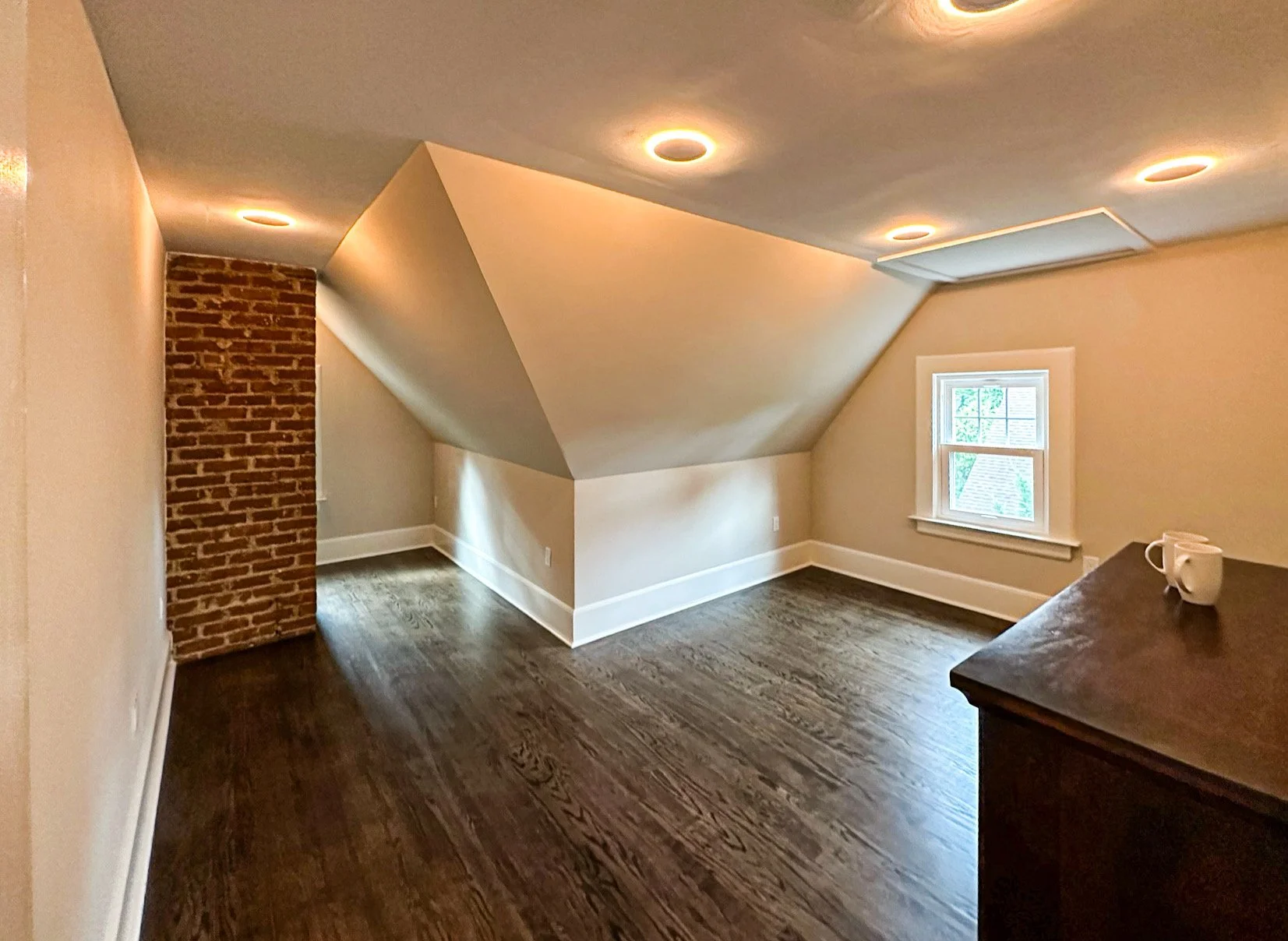Empty attic room with sloped ceilings, a brick column, a small window, recessed lighting, and dark hardwood floors.