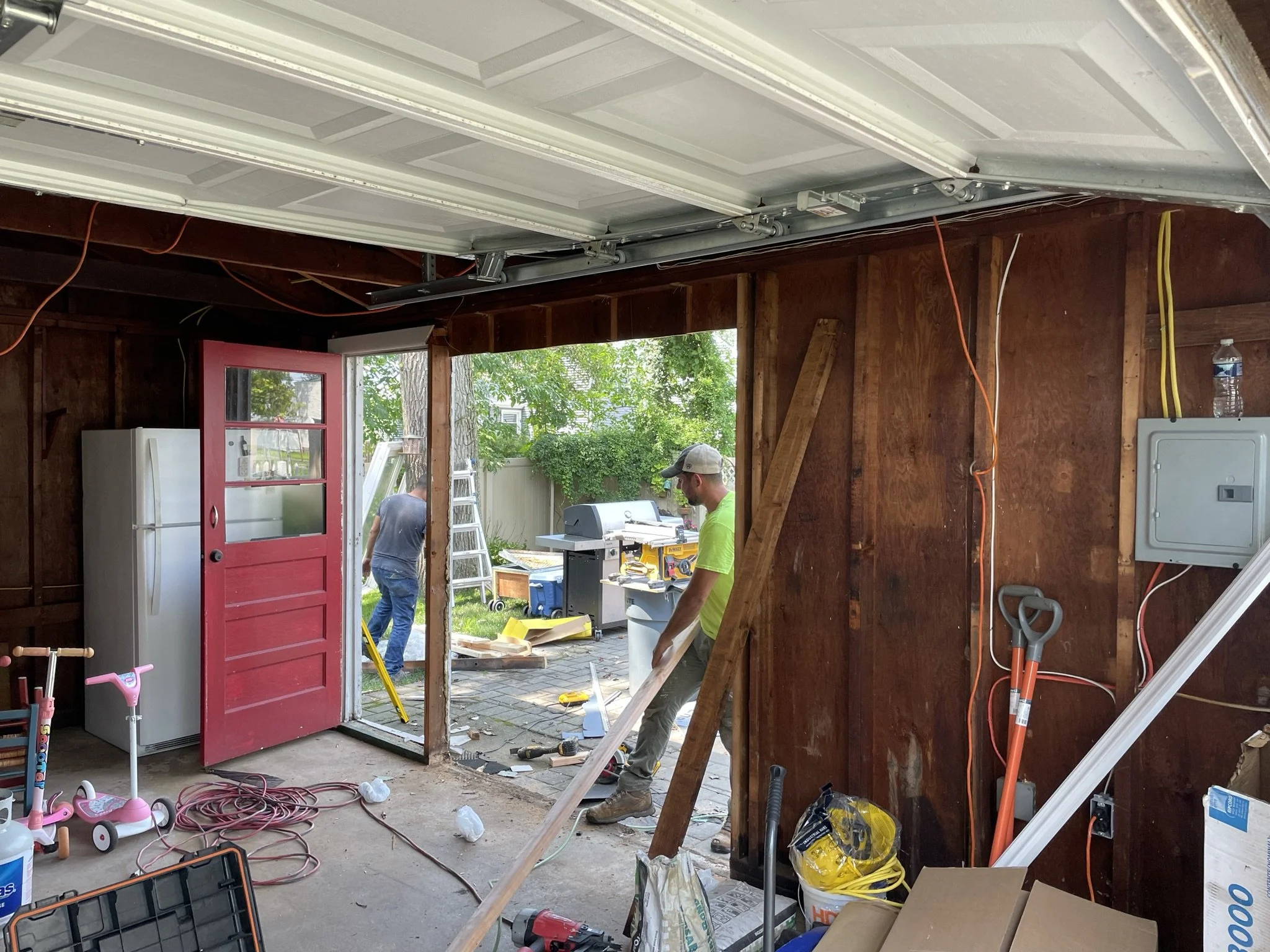 Inside a garage during renovation with two workers outside working on the yard, tools and supplies scattered around.