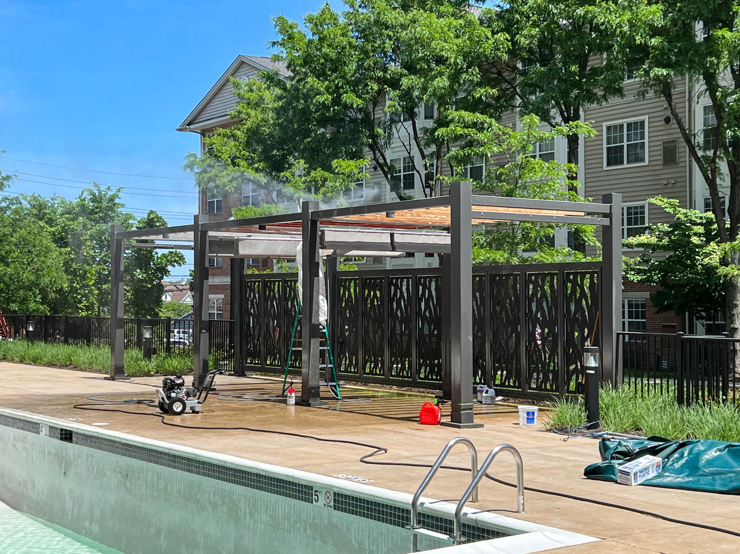 A swimming pool deck with construction or maintenance underway, including cleaning equipment, tools, and materials, surrounded by residential buildings and green trees.
