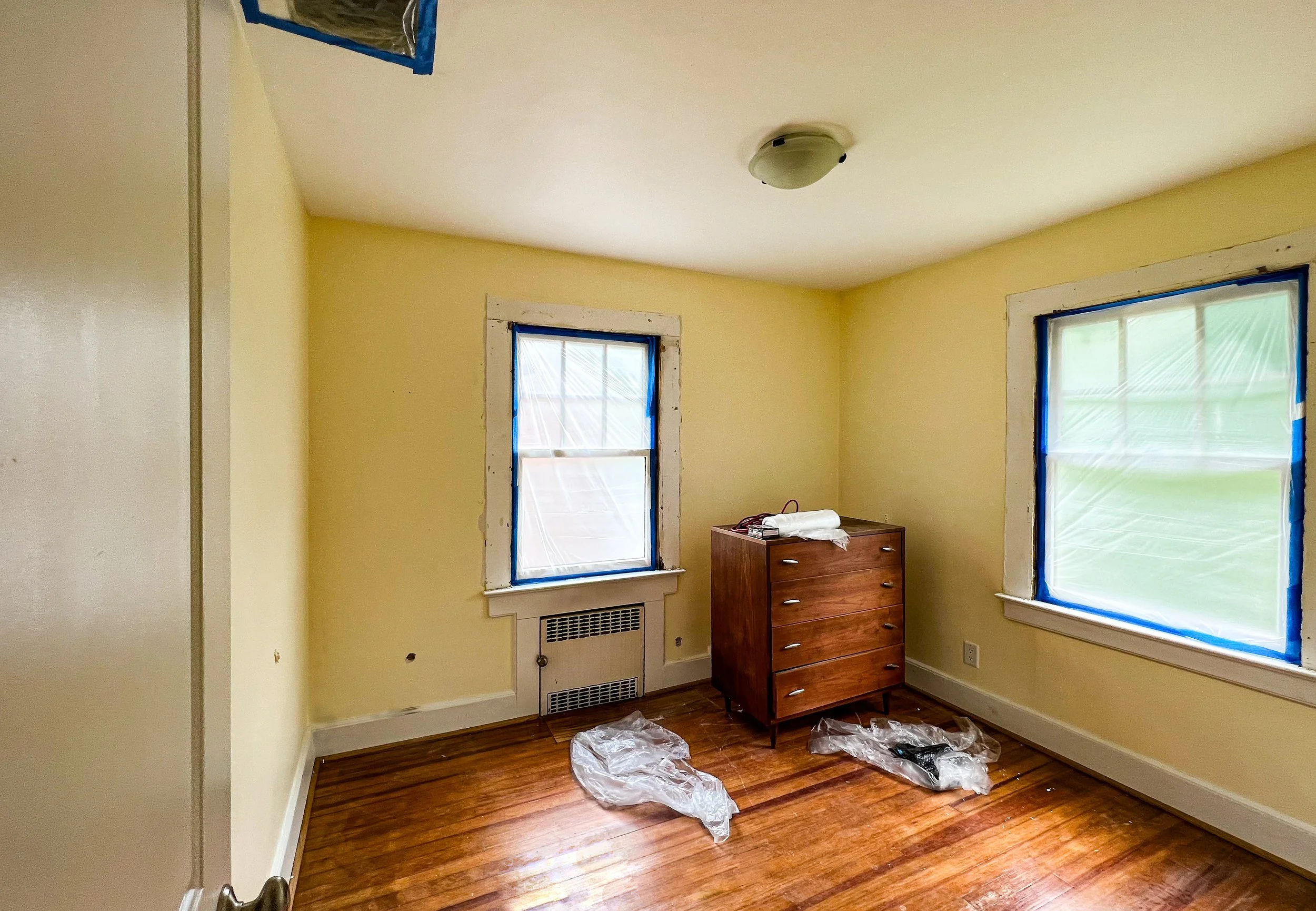 Empty room with yellow walls, hardwood floor, two windows covered with blue painter's tape, a small wooden dresser, and plastic bags on the floor.
