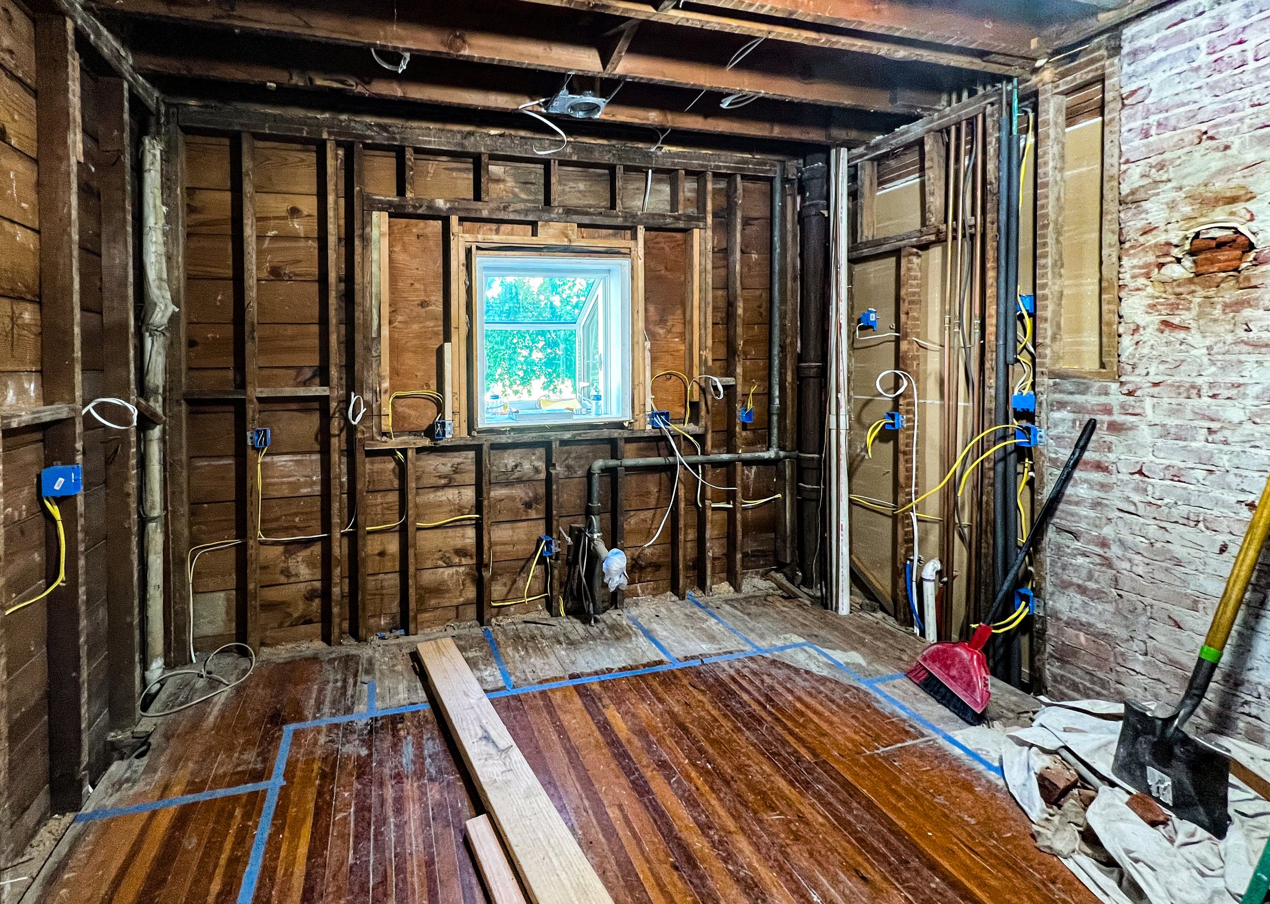 Room under renovation with exposed wooden studs, electrical wiring, and a window. Construction tools and materials, including a broom, shovel, and wooden planks, are present.