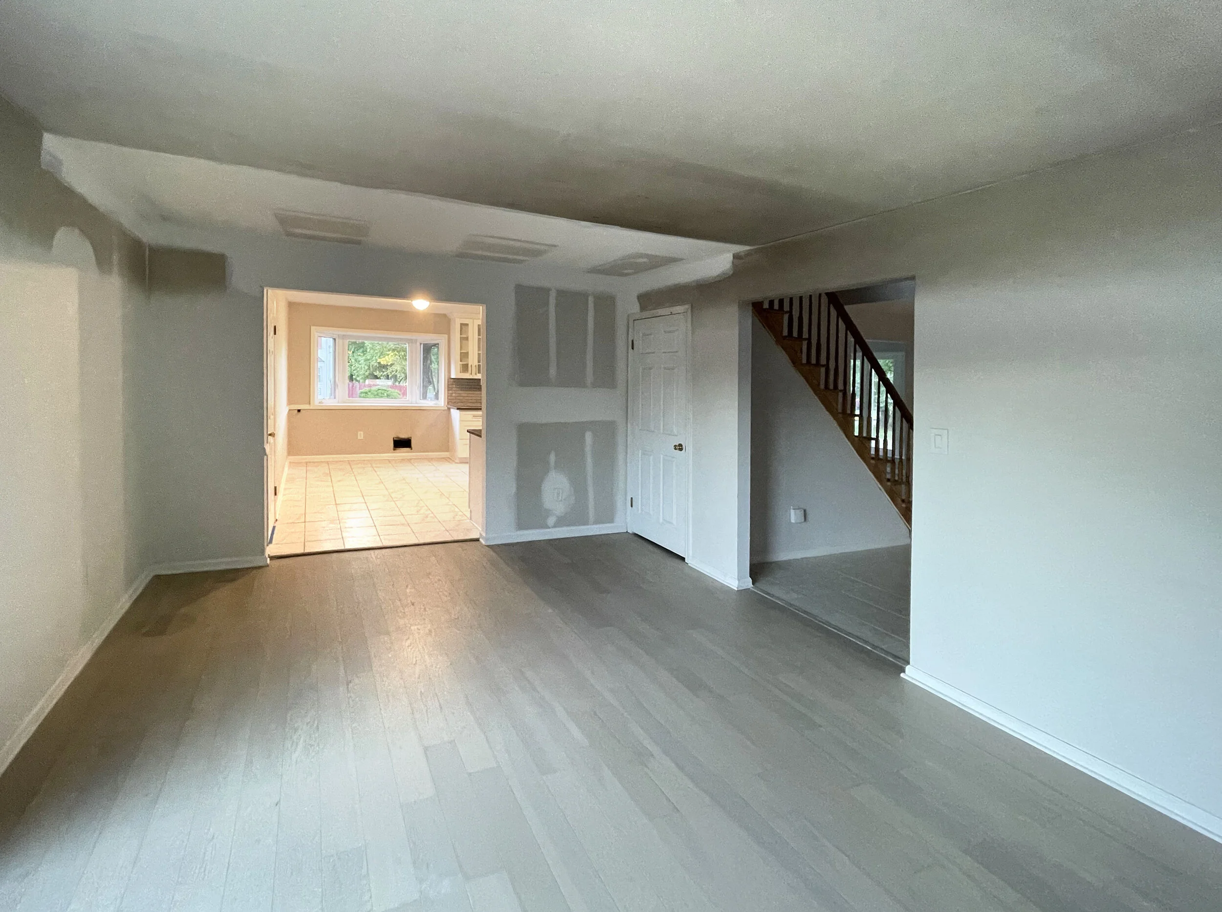 Empty living room with new gray hardwood floors, an open doorway to a kitchen with tiled floor, staircase with wooden railing, and walls in the process of being painted.