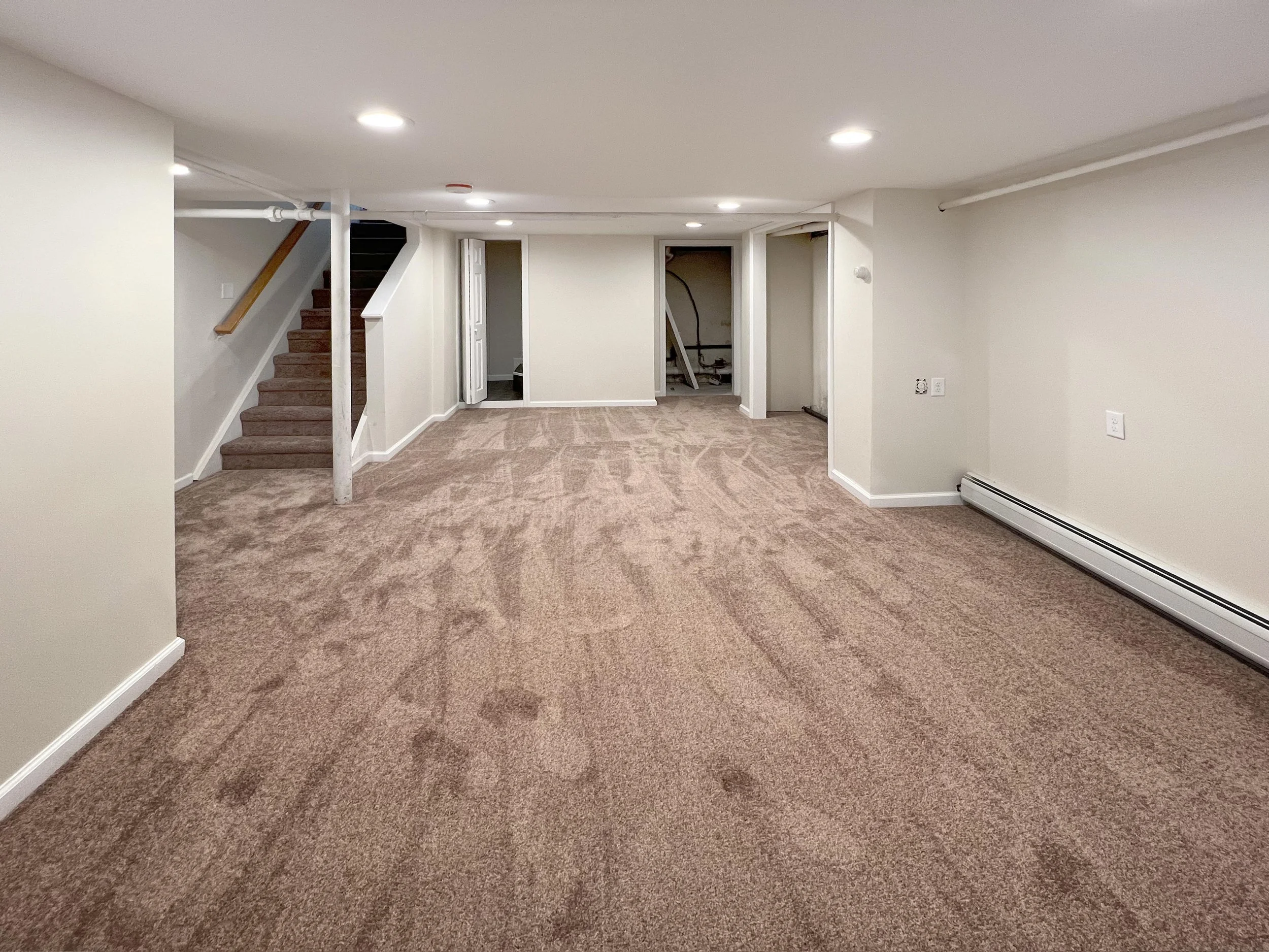 Empty basement room with beige carpet, white walls, and recessed lighting. Staircase on the left leading up, and three doorways in the back wall, one open showing pipes and construction.