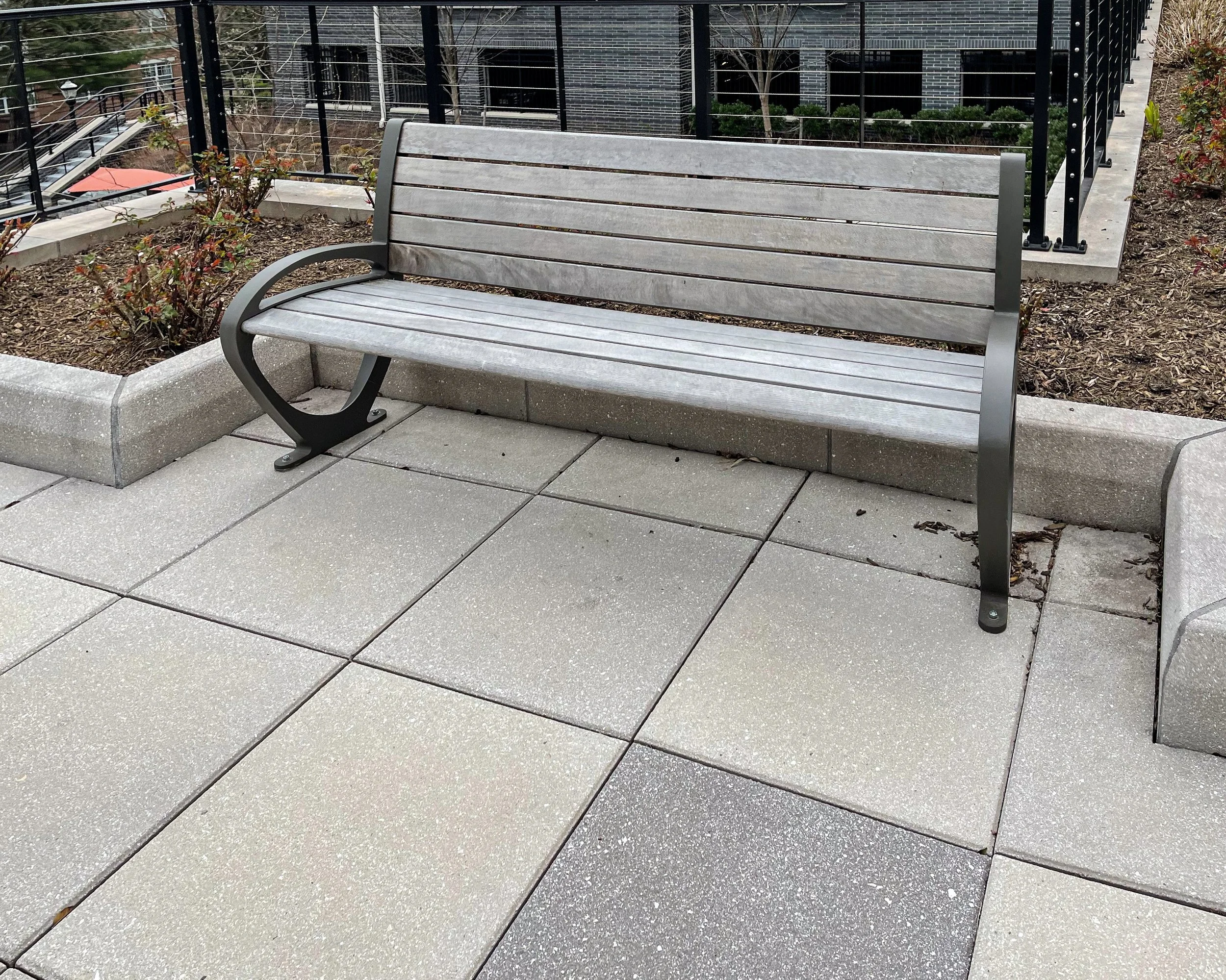 Empty wooden bench on a concrete sidewalk area with planters and a black metal railing behind it.