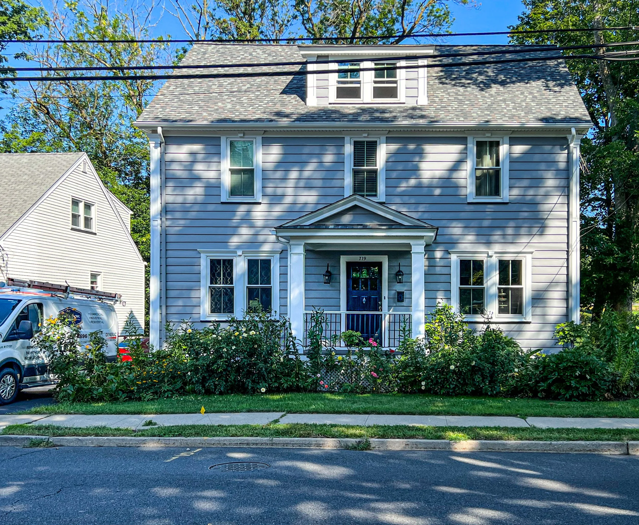 A light blue, three-story house with white trim and a dark blue front door. The house has six windows, three on each of the main floors. There is a small porch with white pillars on either side of the front door, which has two wall-mounted lanterns. 