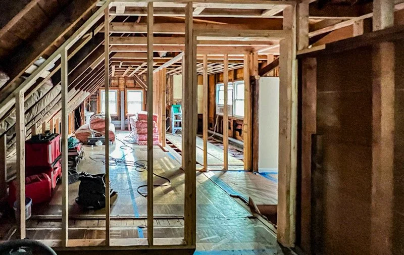 Interior of a house under construction with exposed wooden framing, insulation, and partially installed flooring, with windows at the far end.