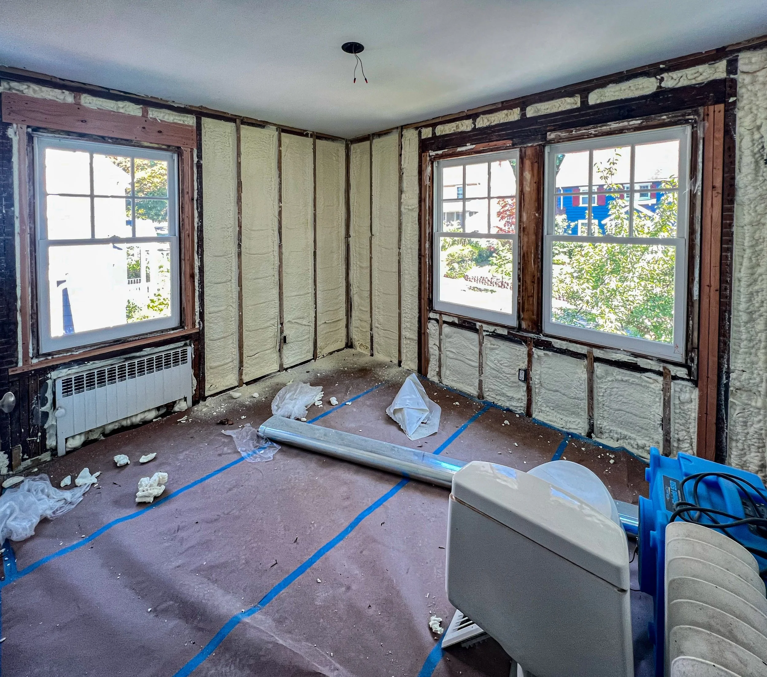 Room under renovation with insulation exposed on the walls, two windows, construction materials on the floor, and a heater below the left window.
