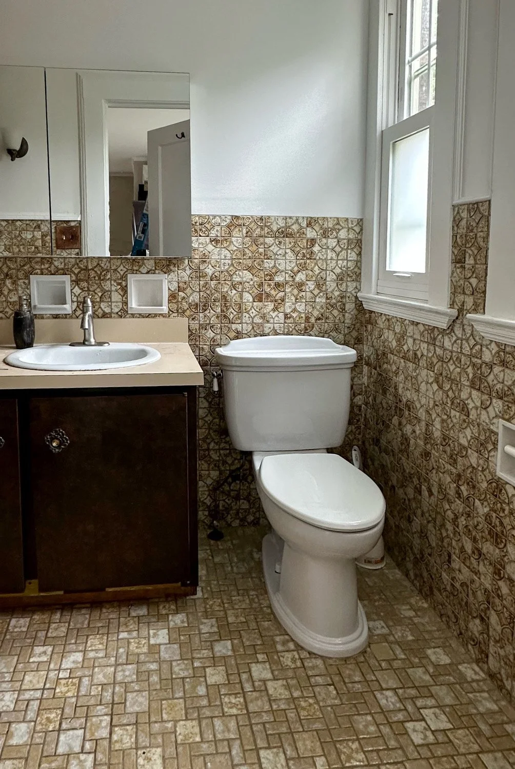 A small bathroom with vintage patterned wall tiles, a white toilet, a beige vanity with a sink and faucet, a wall mirror, a window with white trim, and a tiled floor.