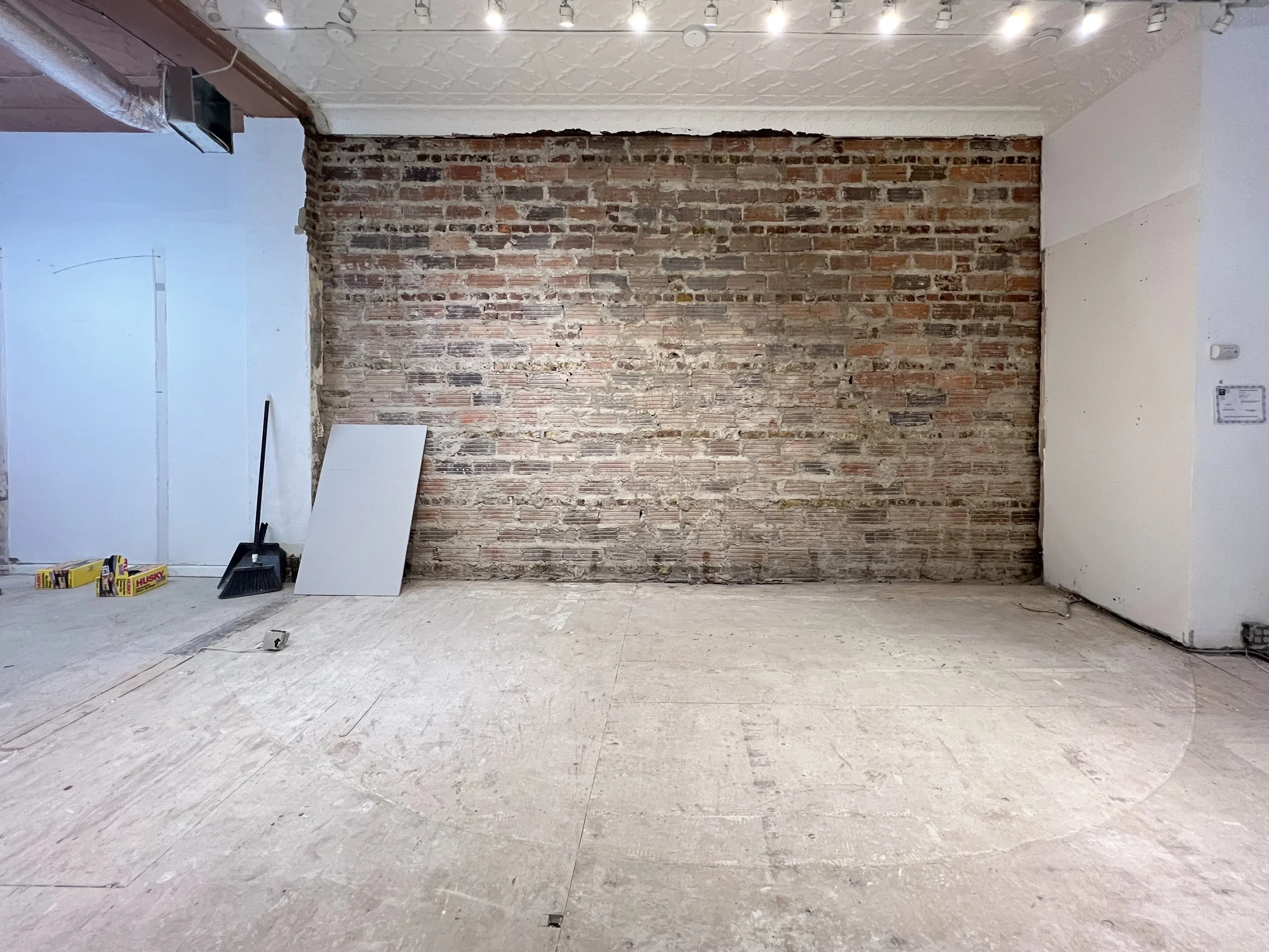 An indoor space under renovation with unfinished flooring, a rustic brick wall, and construction tools including a broom, a white board, and boxes of screws or nails.