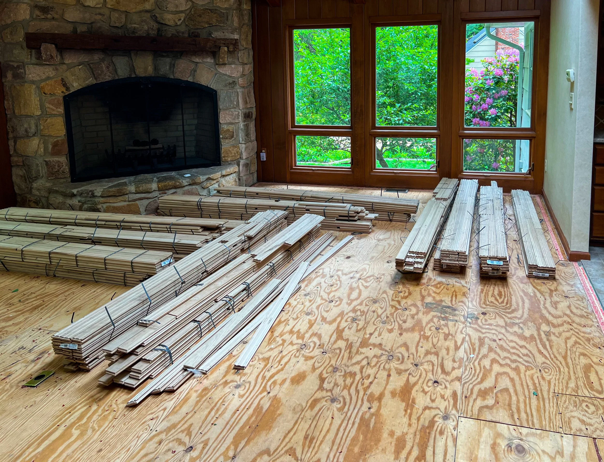 Stacks of wooden flooring planks and molding arranged on a partially installed plywood floor in front of a large window with a view of green trees and pink flowering bushes outside. There is a stone fireplace to the left.