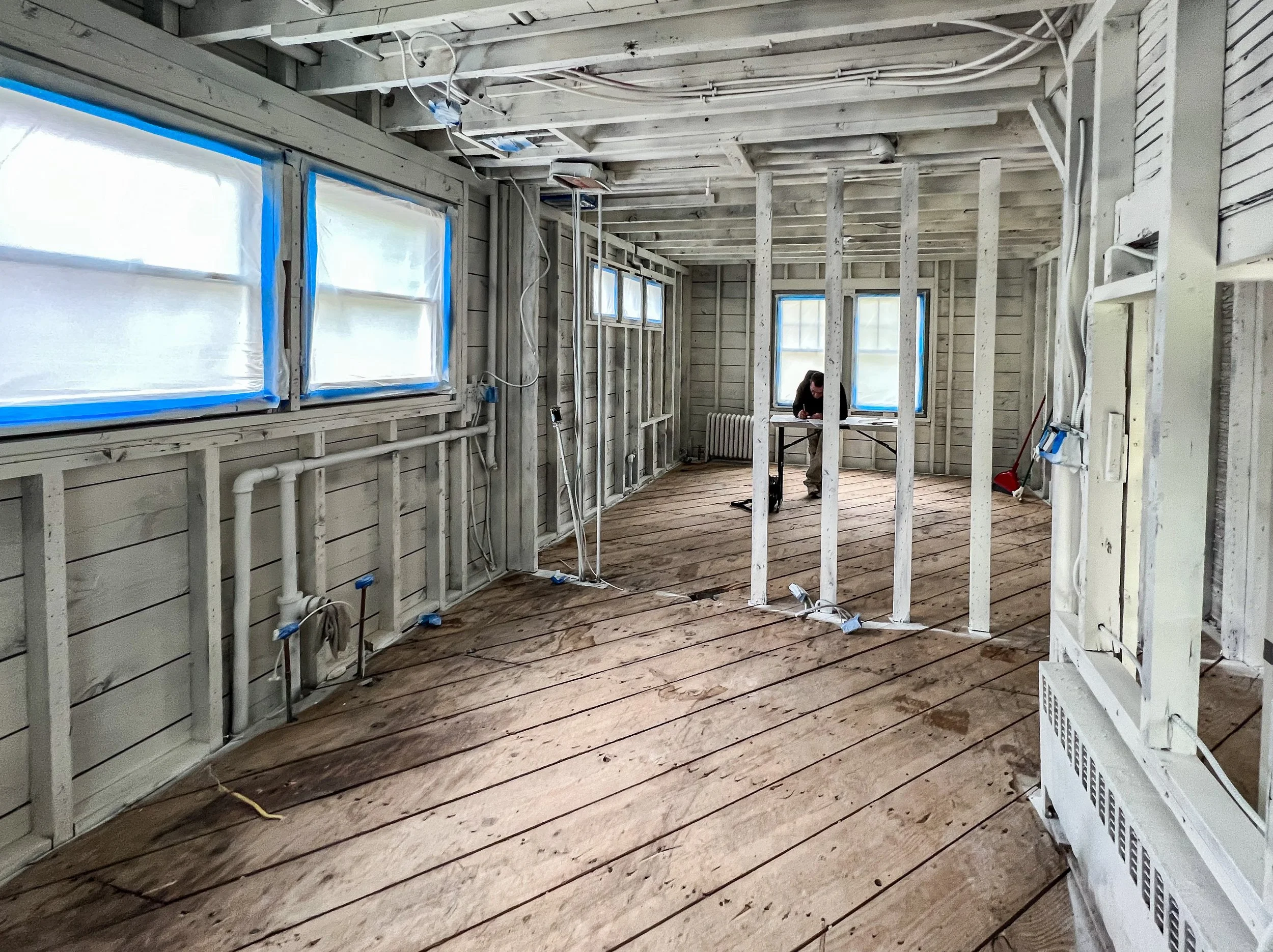 Room under renovation showing wooden floor planks, exposed framing, and windows with blue painter's tape, with a person working at a table.