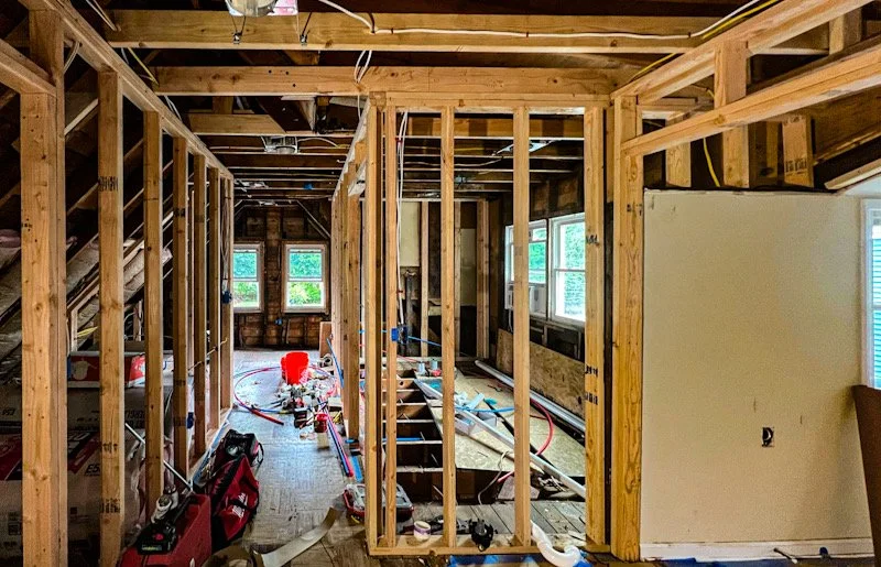 Interior of a house under construction, showing exposed wooden framing, electrical wiring, construction tools and materials, and windows with green landscape outside.