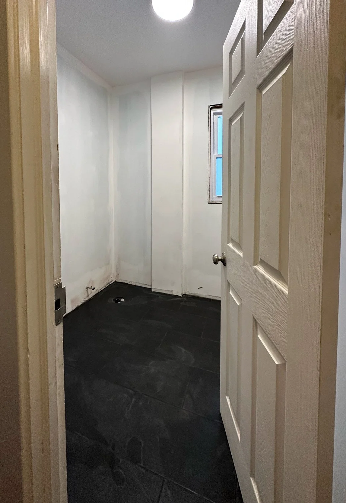 A small, empty room with white walls and a black tiled floor, viewed through an open door with a doorknob. There is a small window on one wall and an overhead light on the ceiling.