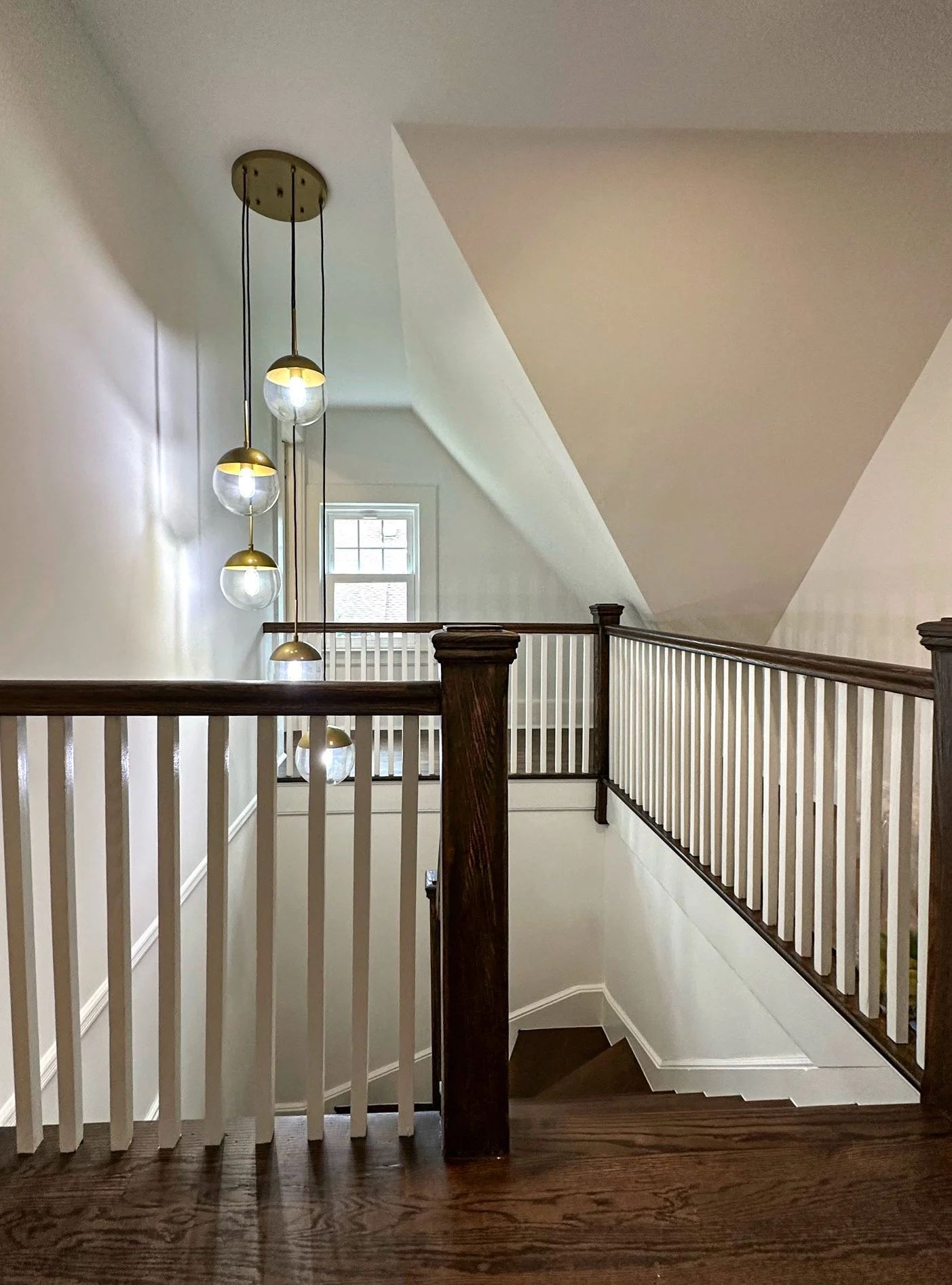 Interior view of a staircase with a wooden handrail and white balusters, a modern pendant light fixture with multiple glass orbs, and a window in the background.